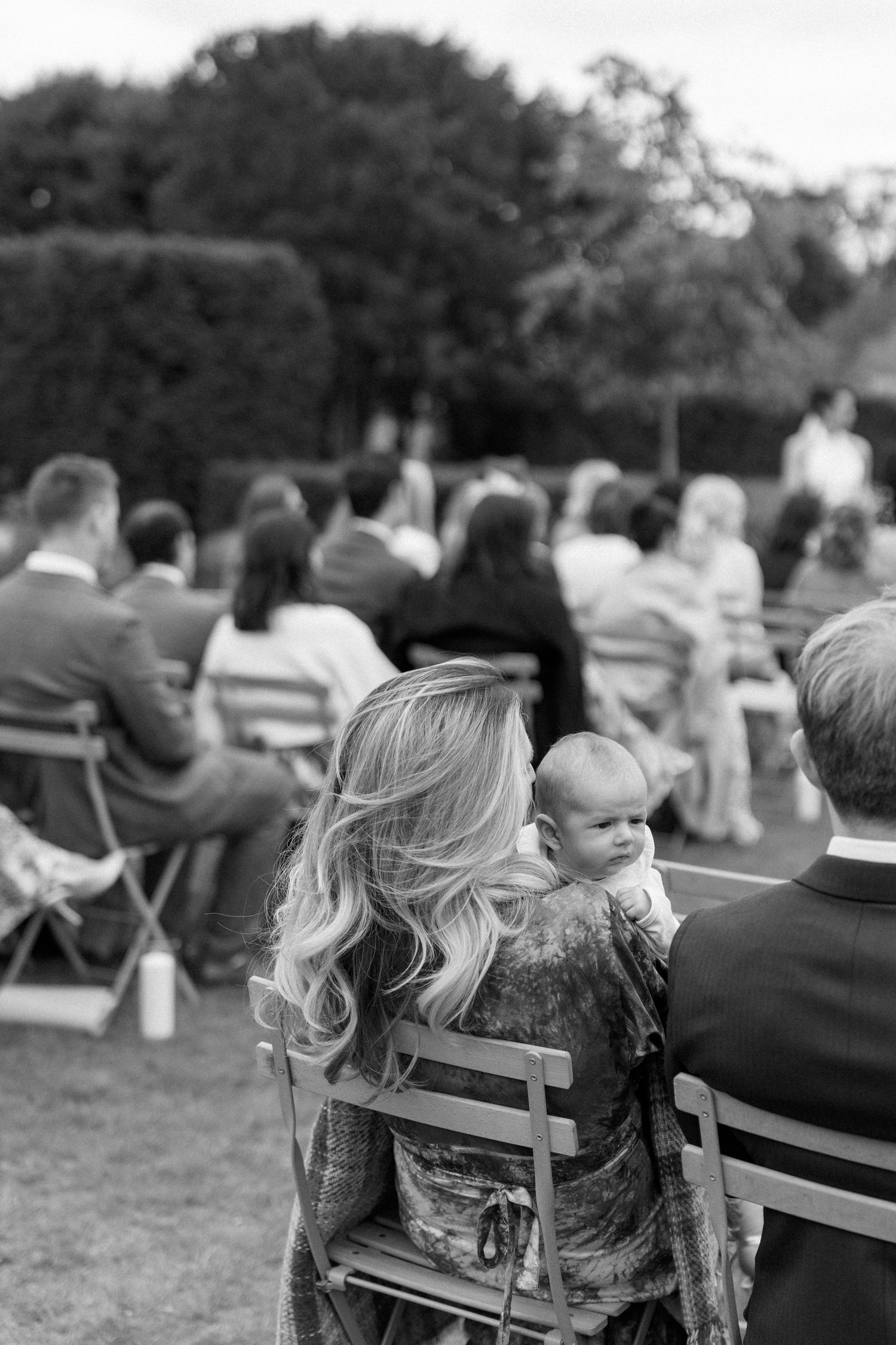Wedding guest holding baby while seated at outdoor garden wedding ceremony.