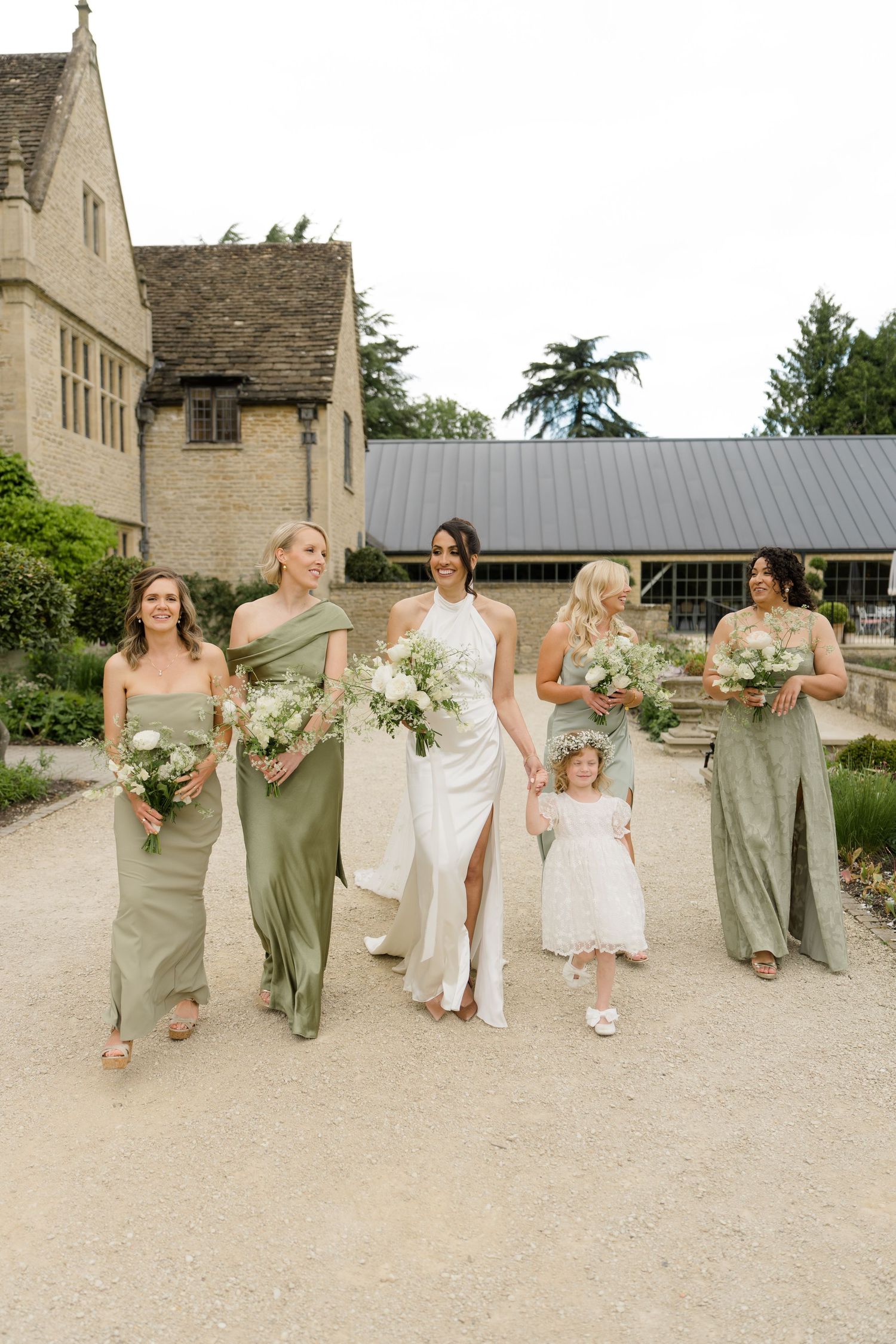 Bride in white halter gown walks with bridesmaids in sage green dresses and flower girl outside stone manor.