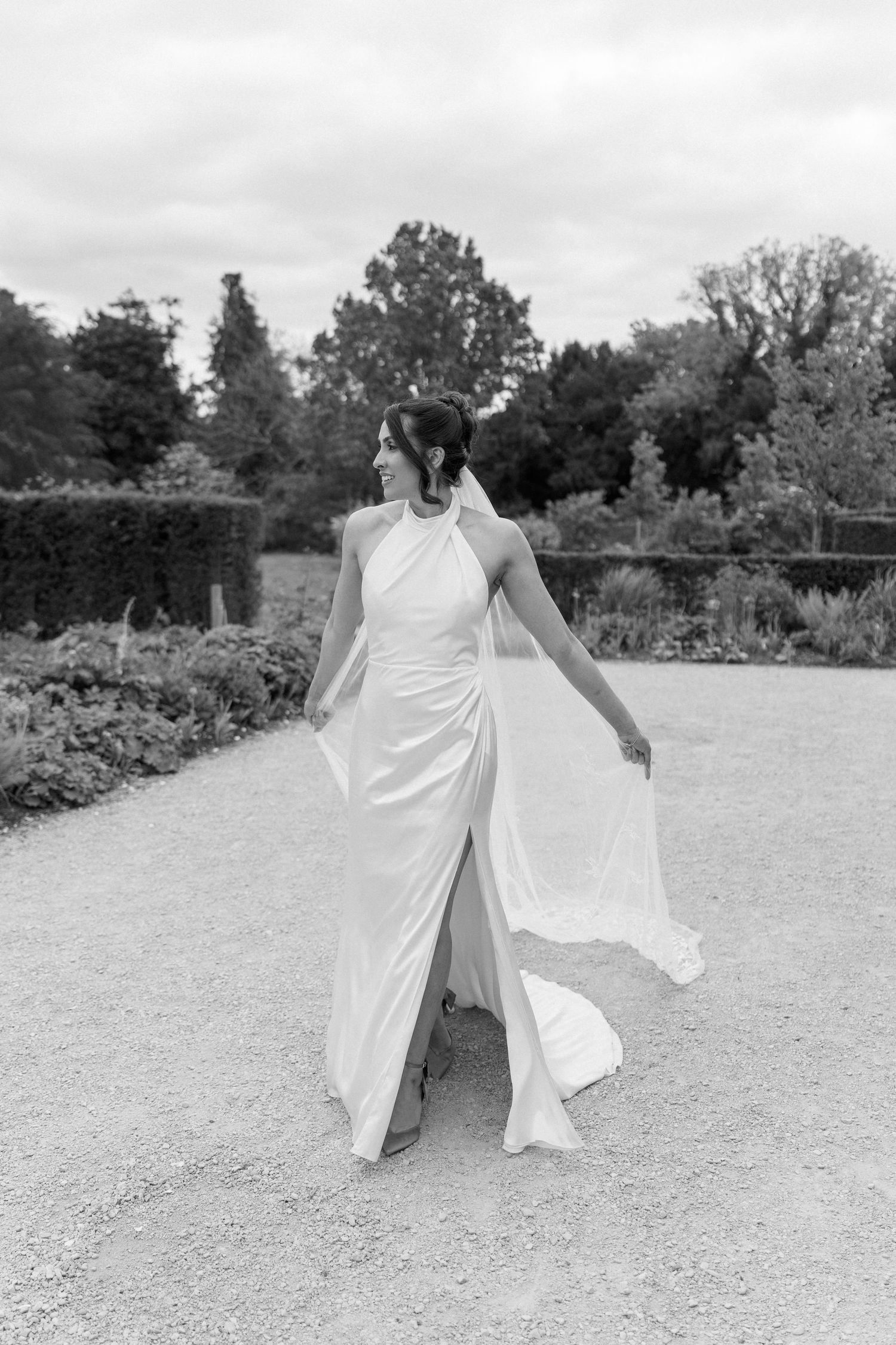 Black and white portrait of bride in elegant halter-neck wedding gown with flowing skirt walking in formal garden.