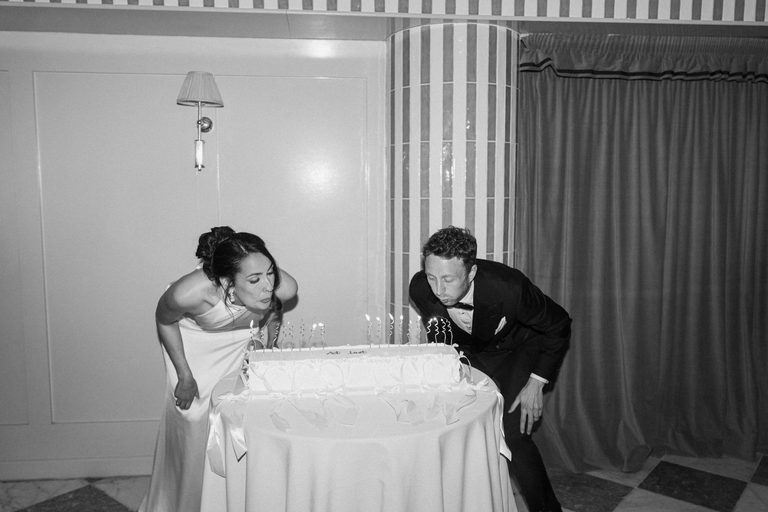 Black and white photo of bride and groom blowing out candles on a wedding cake together.