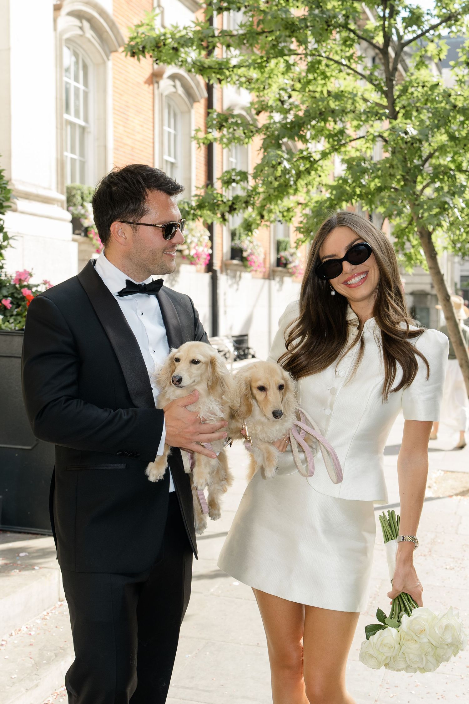 Smiling bride in white mini dress and groom in tuxedo carry two dachshund puppies on sunny street.