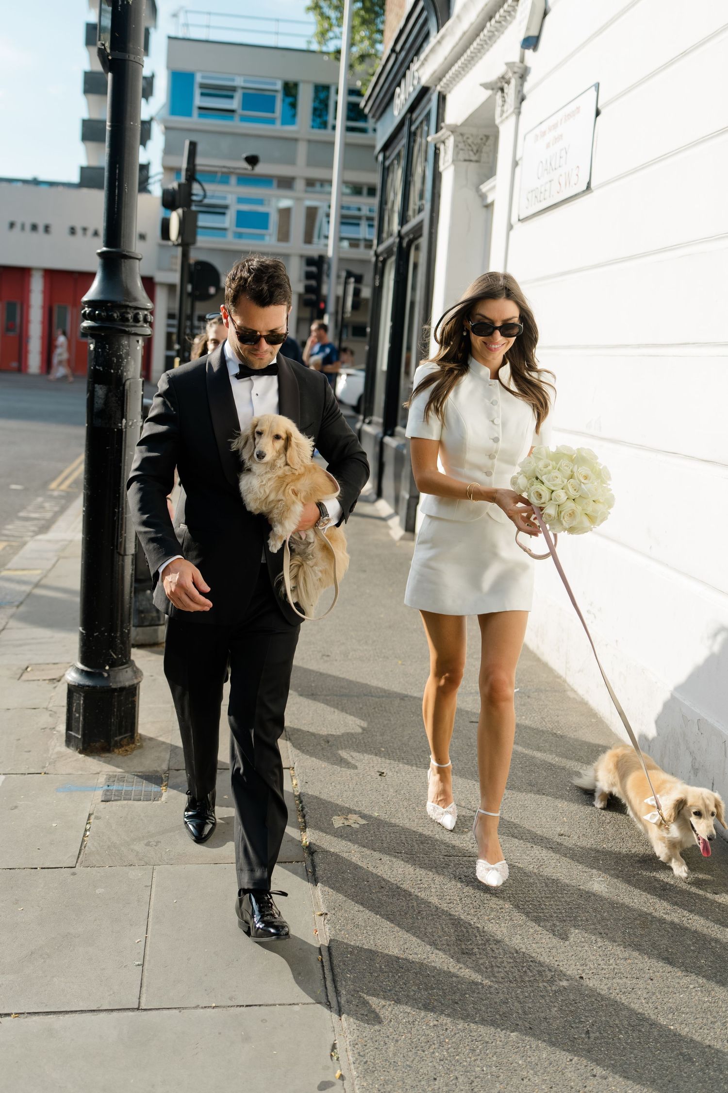 Groom carrying cocker spaniel while bride walks dog on leash in white mini dress.