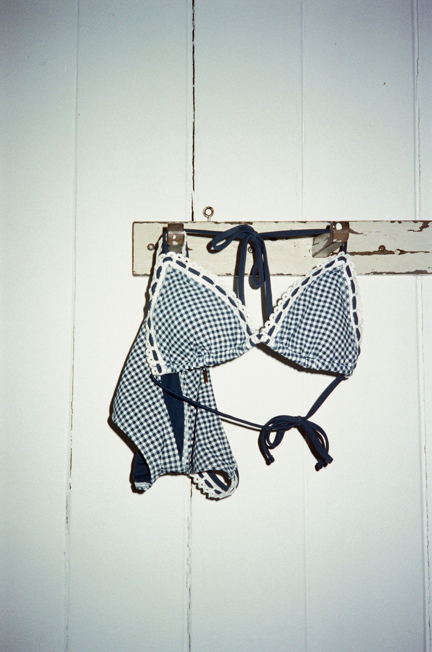 A blue gingham bikini set hangs on a rustic white wooden wall hook.