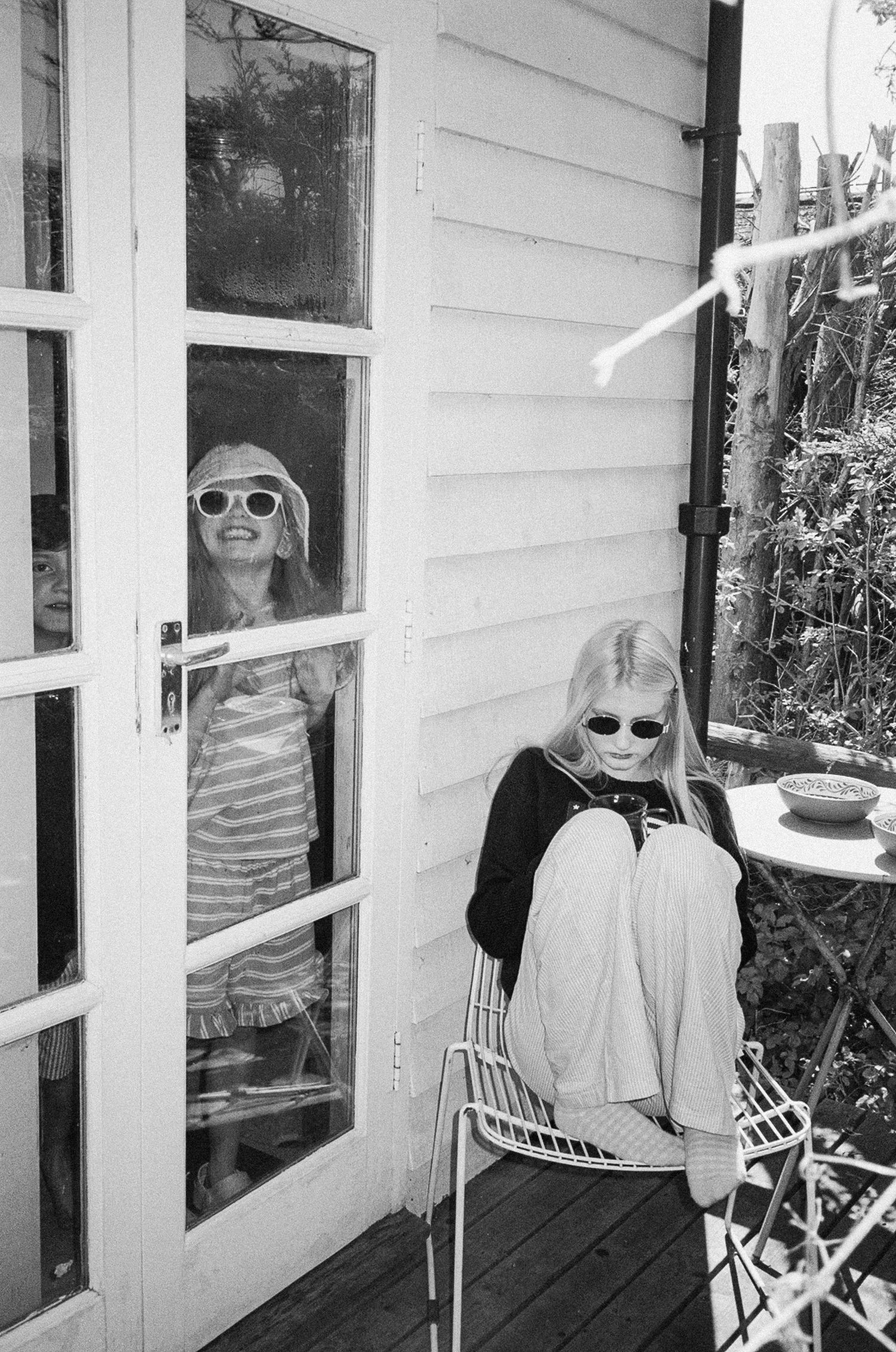 Two blonde girls wearing sunglasses on a porch, one seated in a chair, one visible through glass French doors.