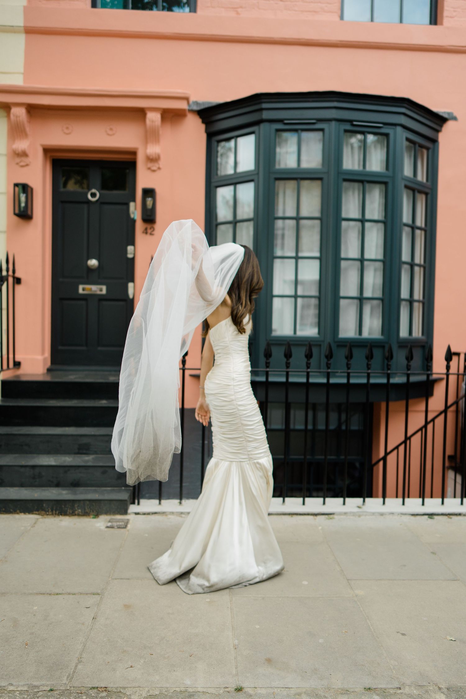 Bride in a fitted white gown and flowing veil stands on a sidewalk outside a pink building with black trim.