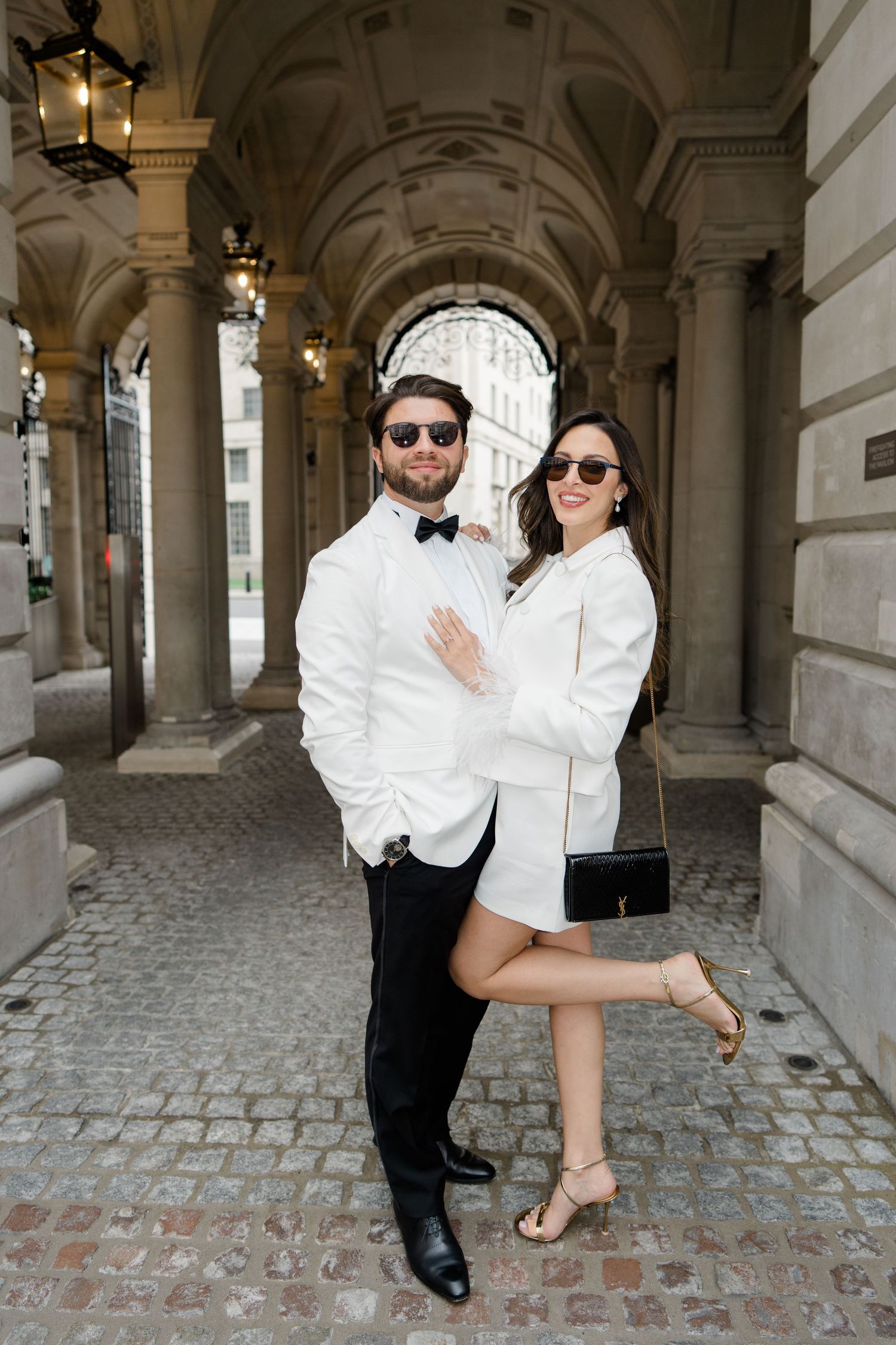 Elegant couple in white outfits posing under stone archway on cobblestone street, wearing sunglasses.