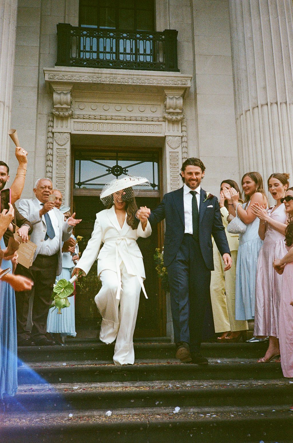 Bride in white suit and hat walks with groom in navy suit down steps as guests celebrate their wedding exit.