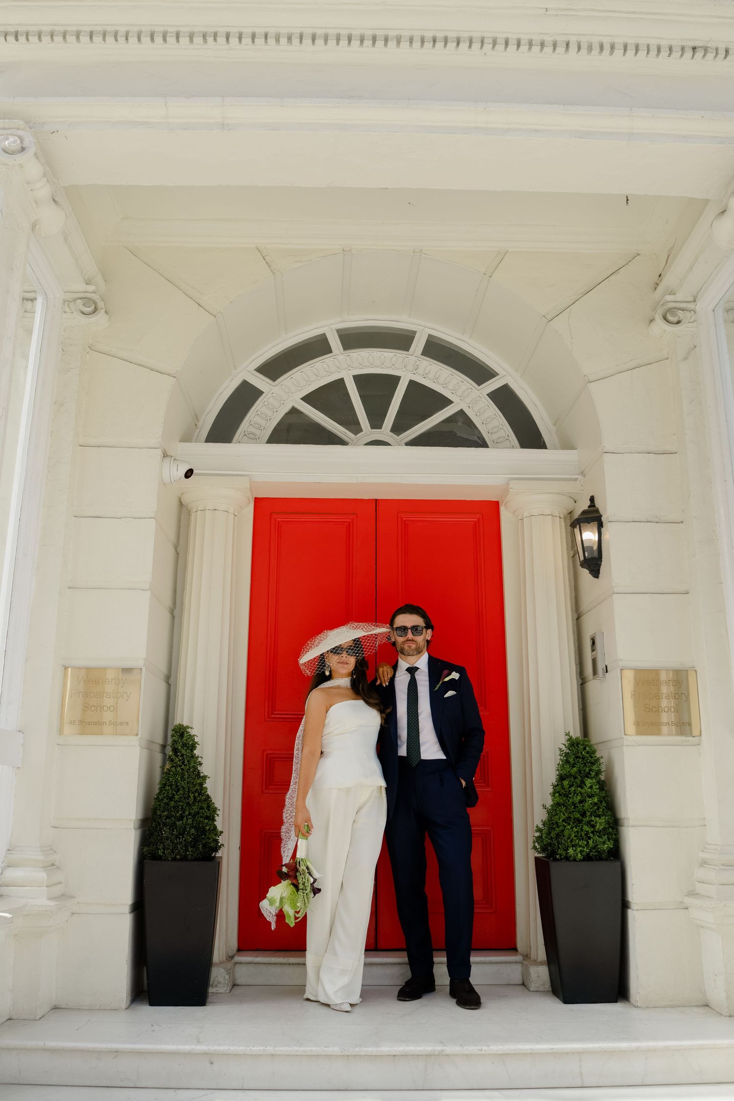 Bride and groom pose before elegant red door of white Georgian building with columns.