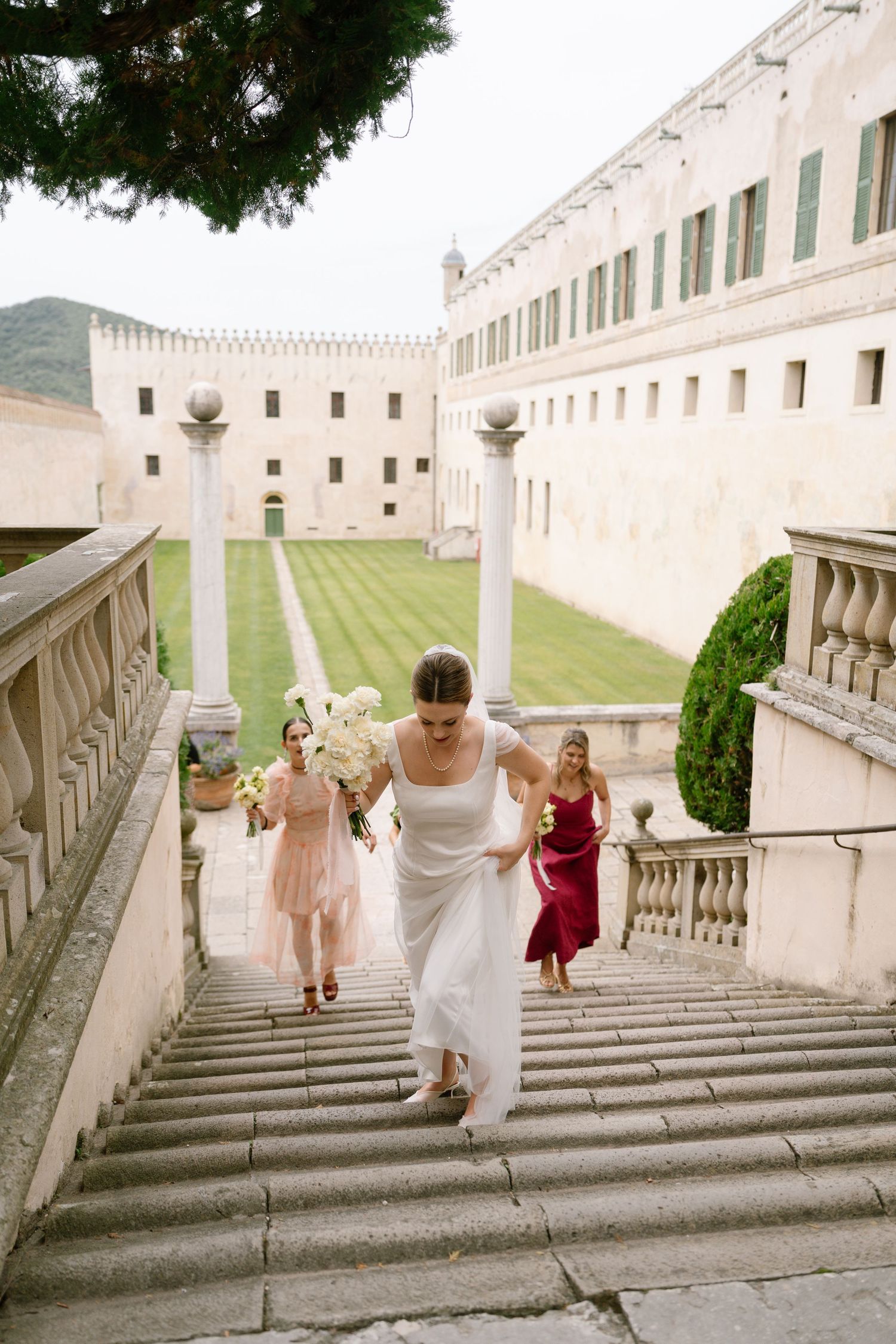 Wedding party descends grand stone steps at a historic venue with manicured lawns.