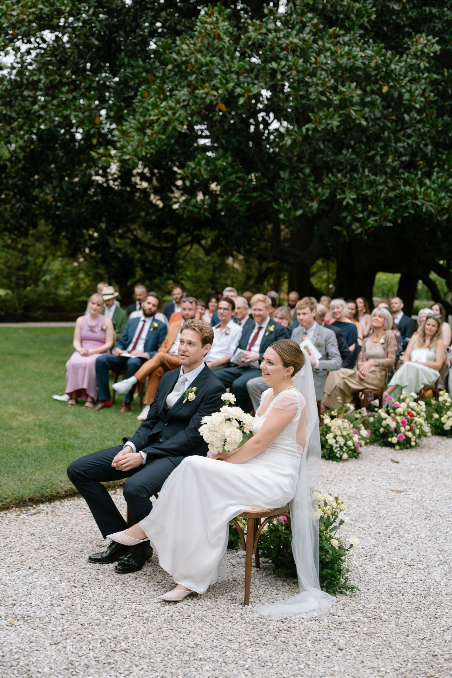 Wedding guests seated on wooden chairs along a gravel path for an outdoor ceremony.
