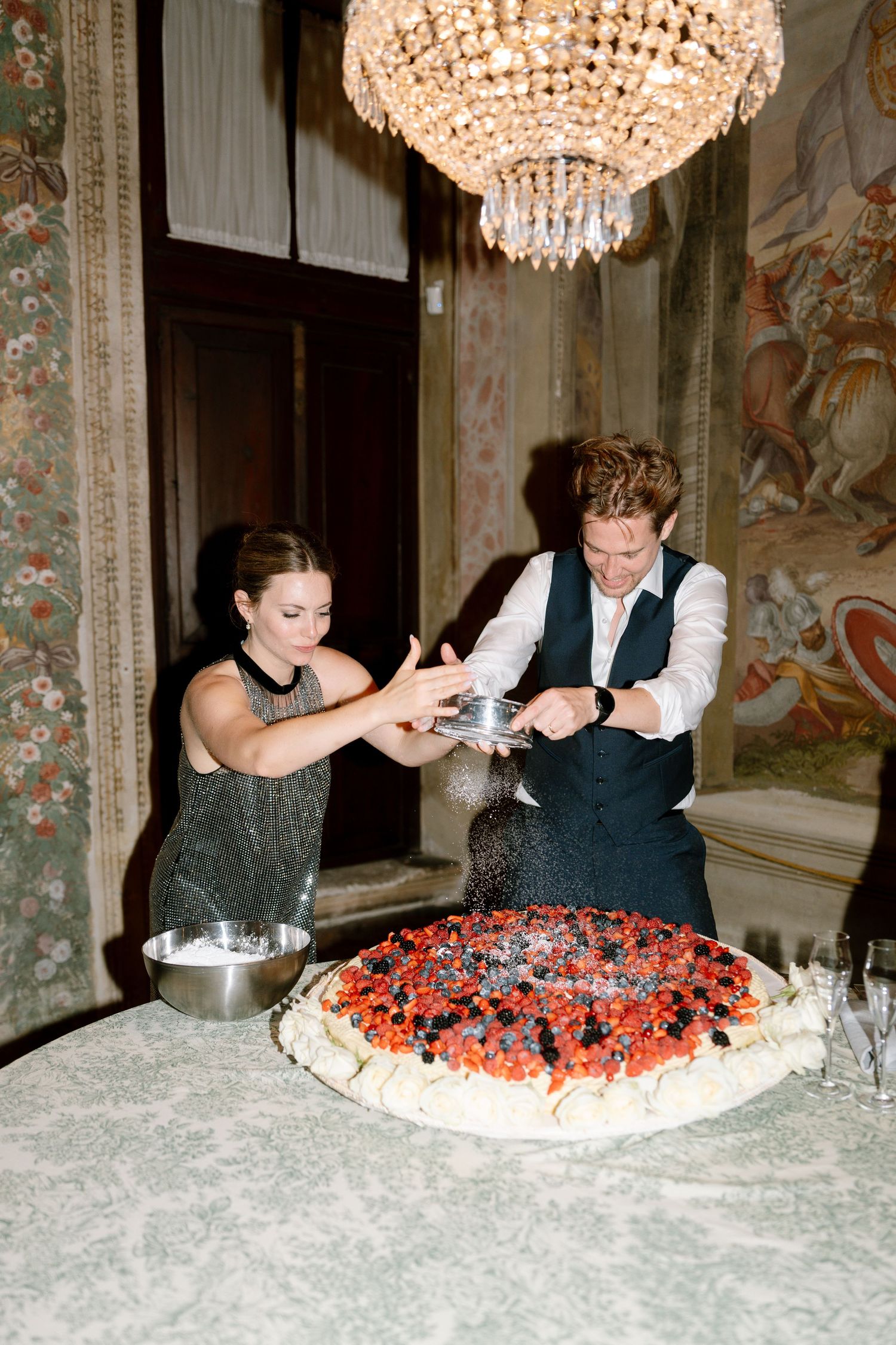 A couple cuts into a large decorated cake beneath a crystal chandelier at an elegant event.