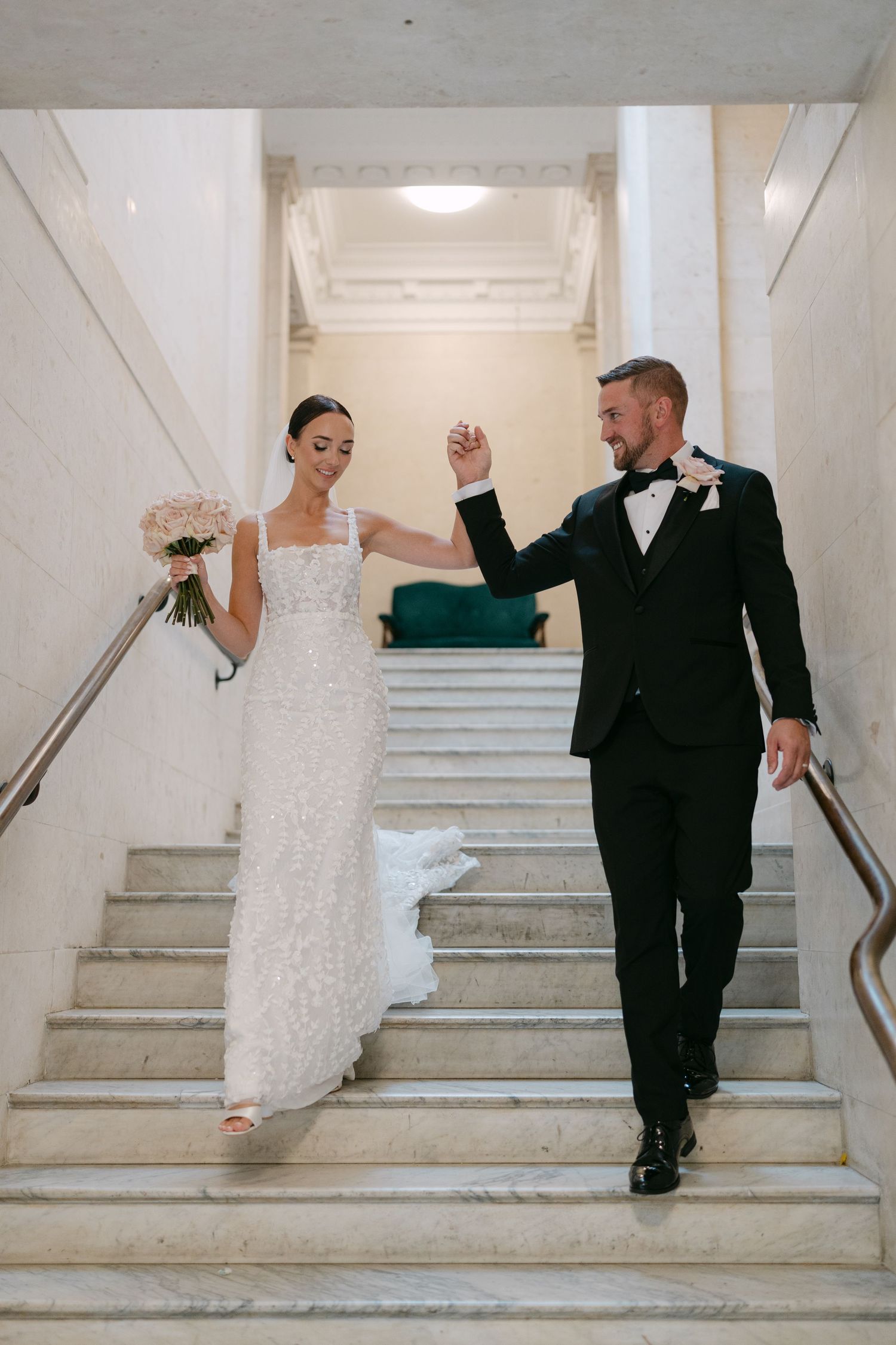 Newlyweds descend white marble staircase holding hands with bride carrying bouquet.