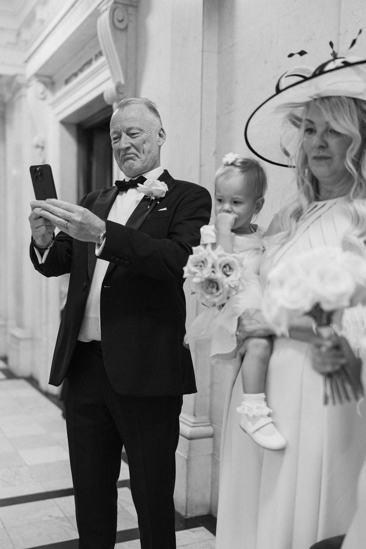 Wedding guests in formal attire gather in an elegant hallway while someone checks their phone.