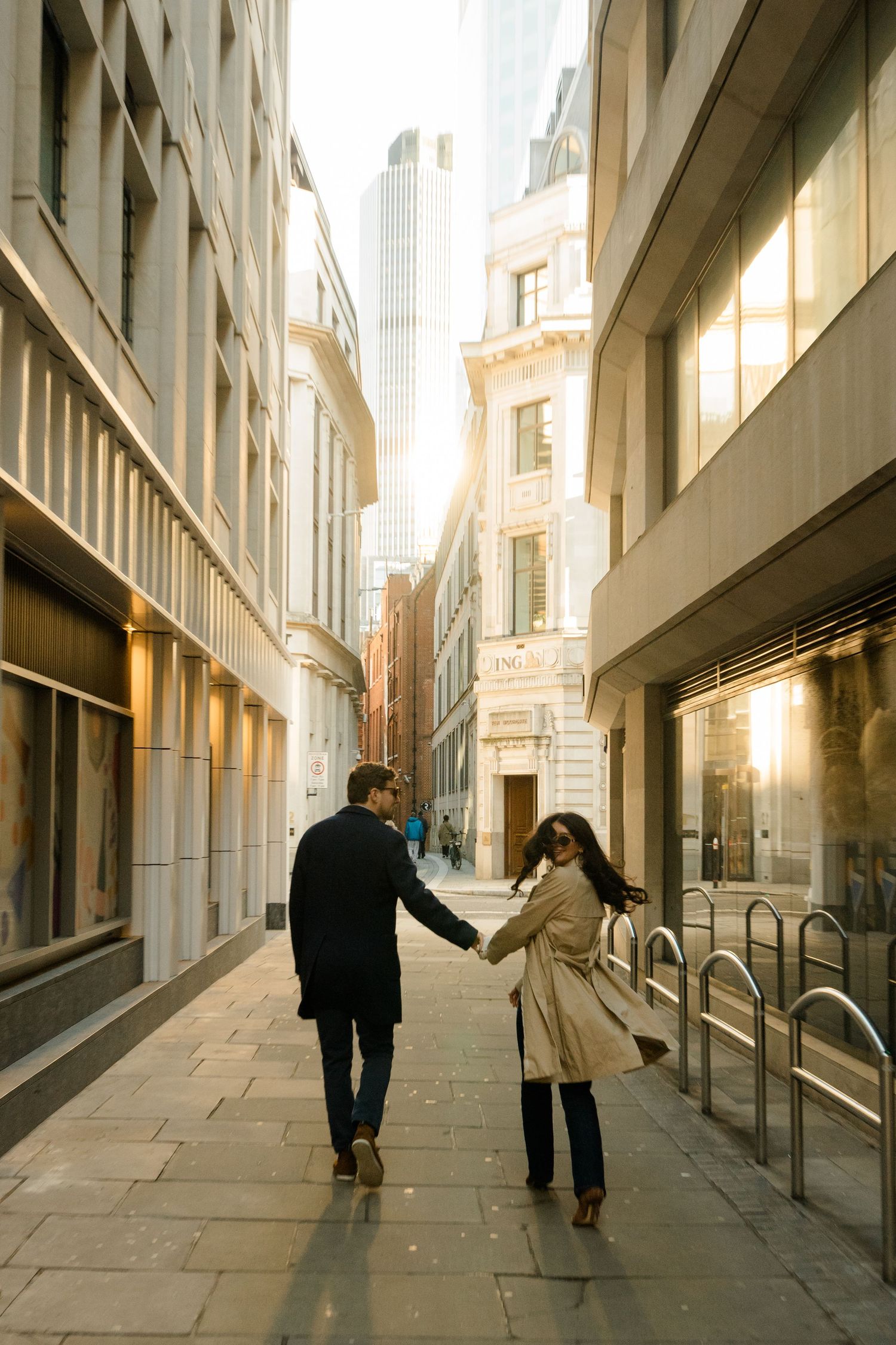 Couple walking through modern urban corridor with tall buildings and dramatic lighting.