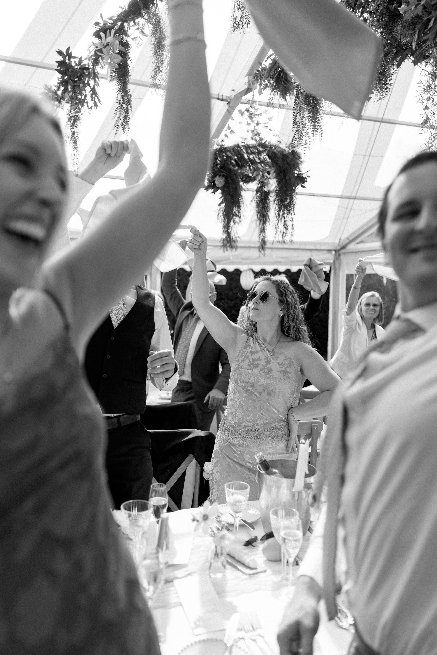 Wedding guests celebrating with raised arms under tent with hanging greenery.
