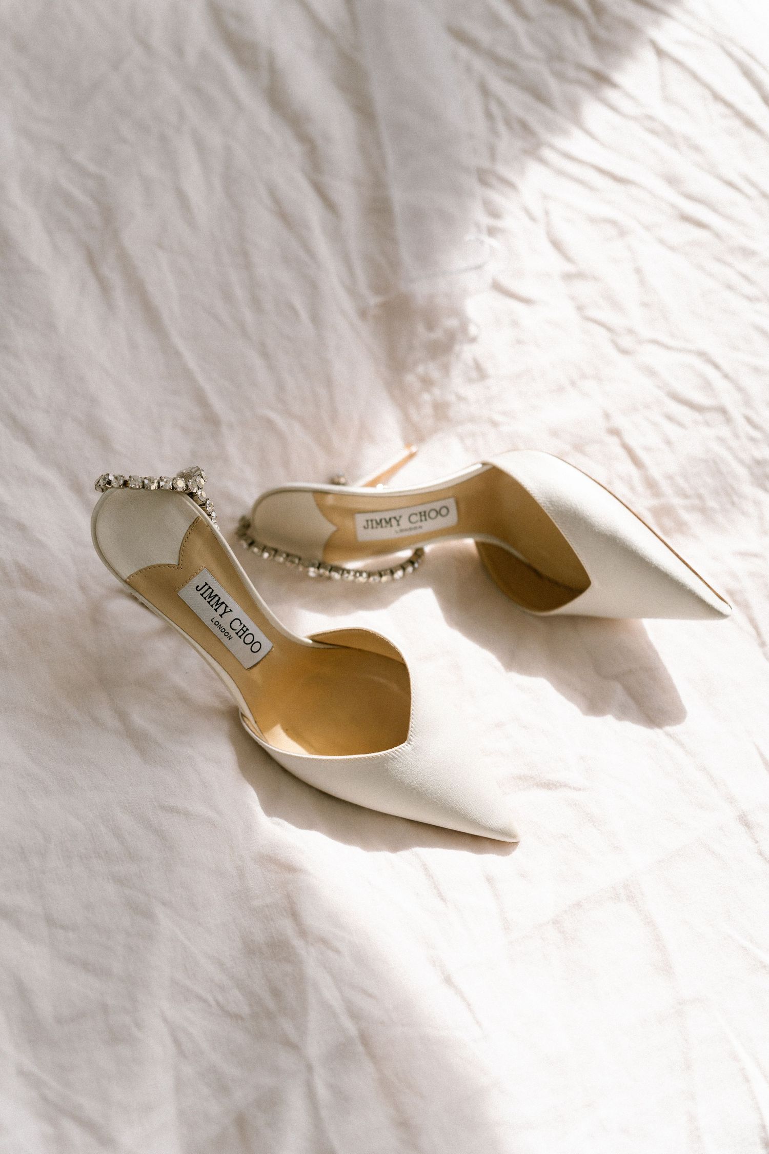 Elegant white Jimmy Choo bridal shoes with crystal embellishments displayed on white fabric.