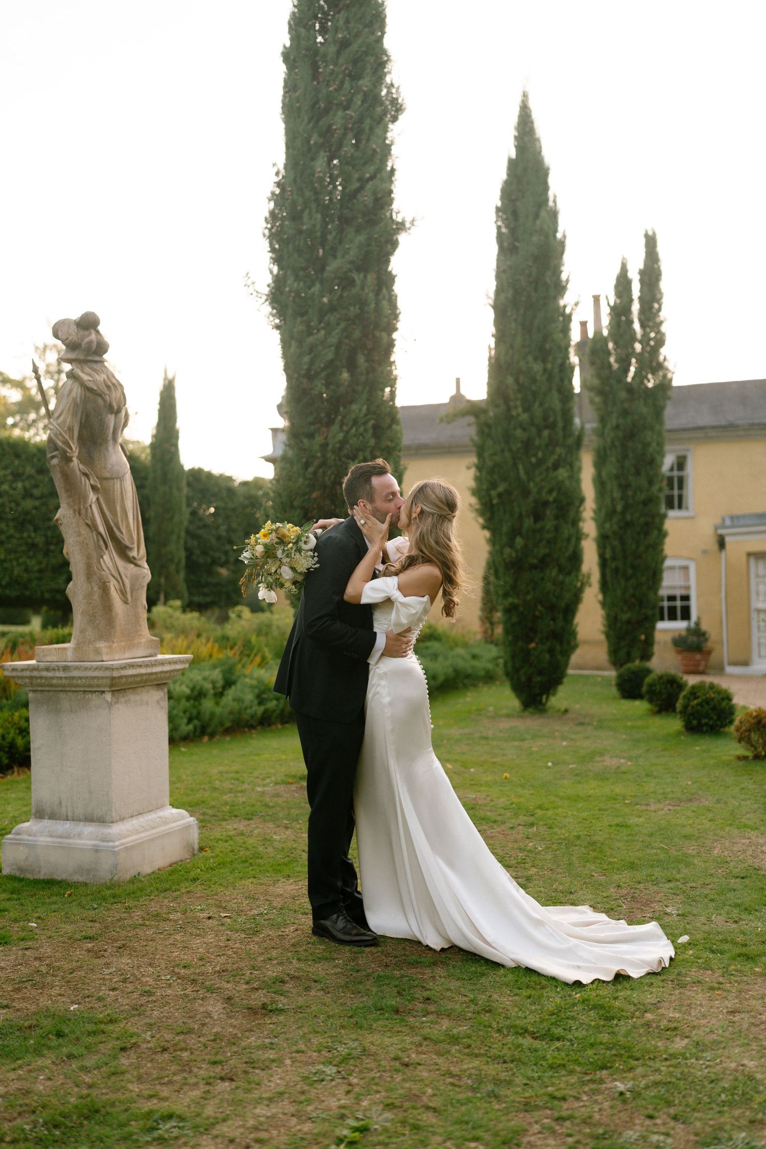 A romantic wedding portrait in a garden with tall cypress trees and classical statuary.