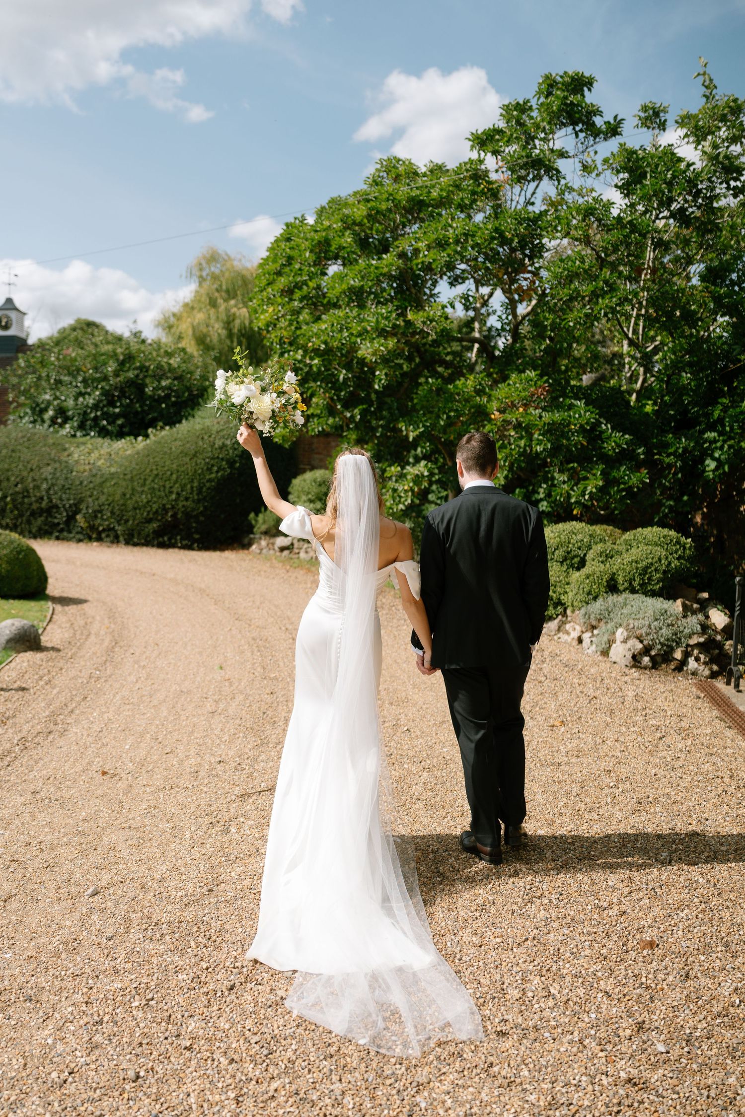 Couple walking down a gravel path in formal attire surrounded by manicured gardens.