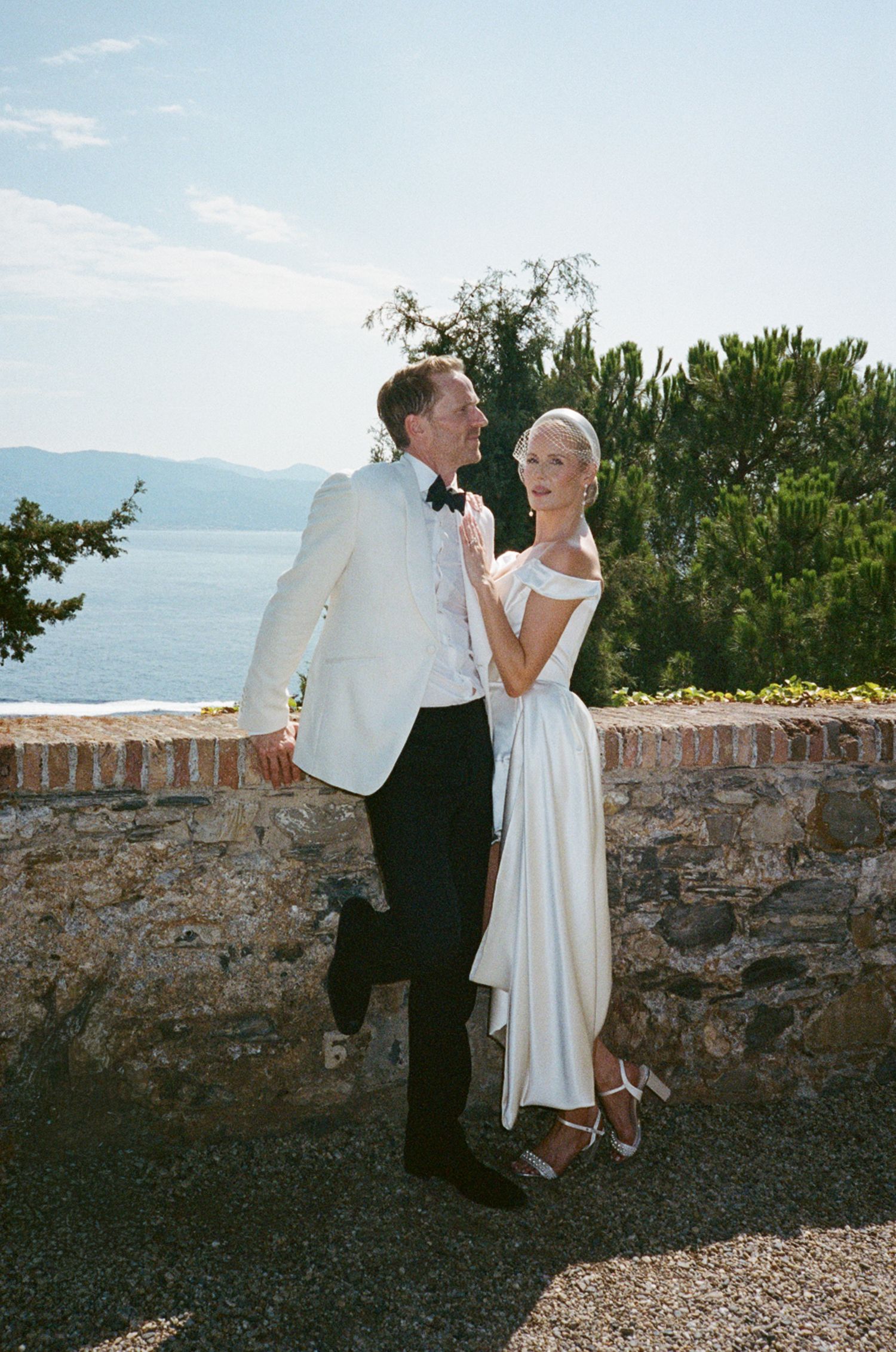 Elegant couple posing outdoors in wedding attire with scenic ocean and mountain backdrop on a sunny day.