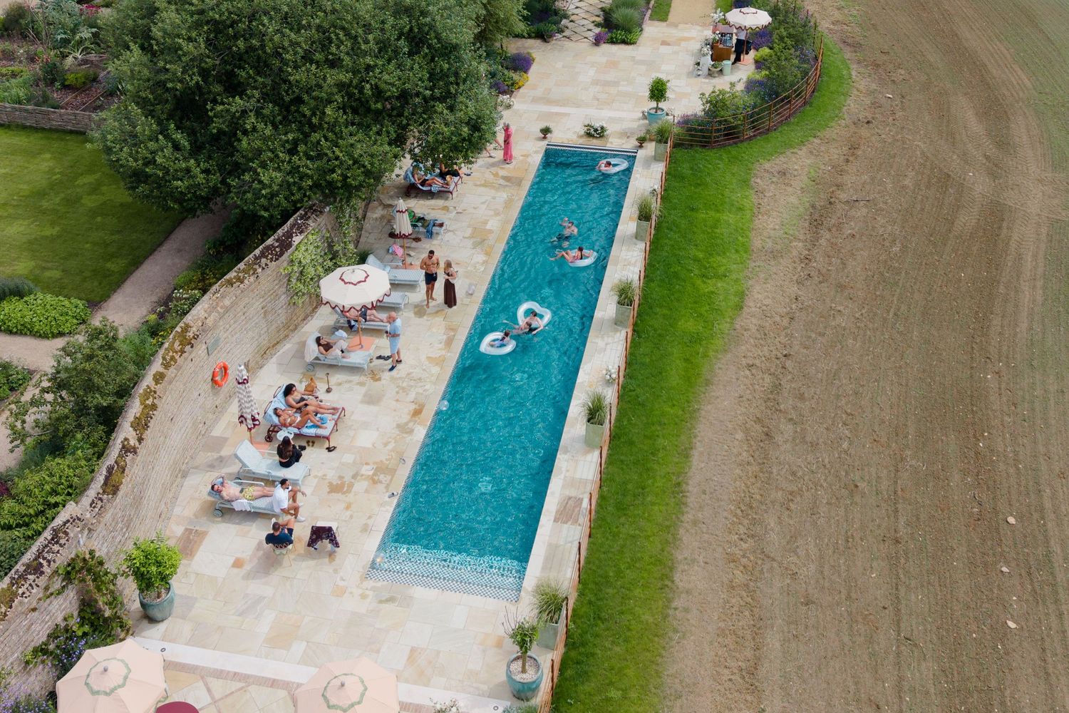 Aerial view of narrow rectangular swimming pool with guests relaxing on loungers beside manicured garden.