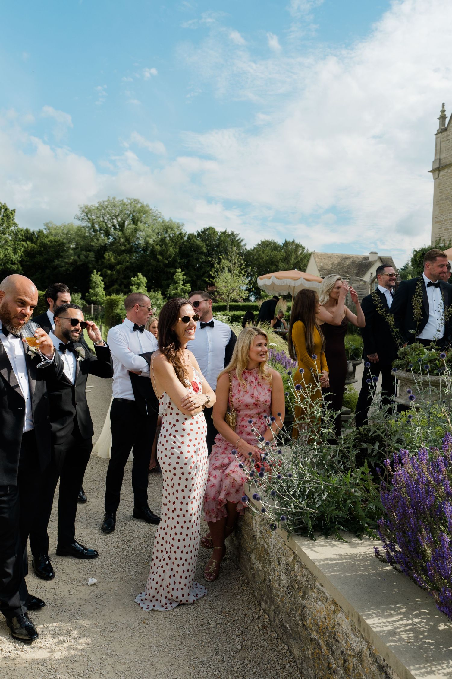Wedding guests in formal attire socializing outdoors in a sunny garden with blue skies above.