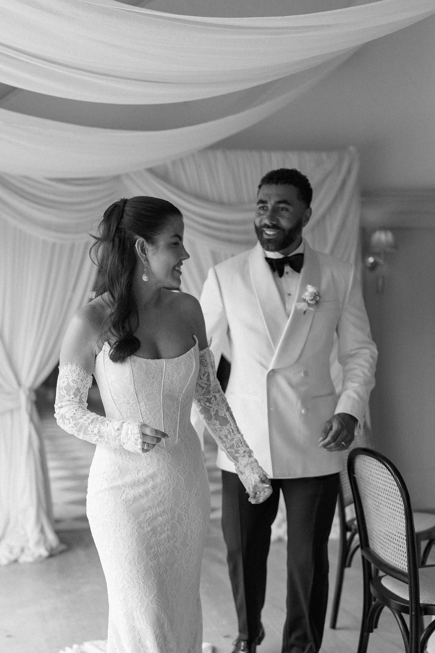 Black and white photo of smiling bride and groom in white tuxedo walking together under draped fabric.