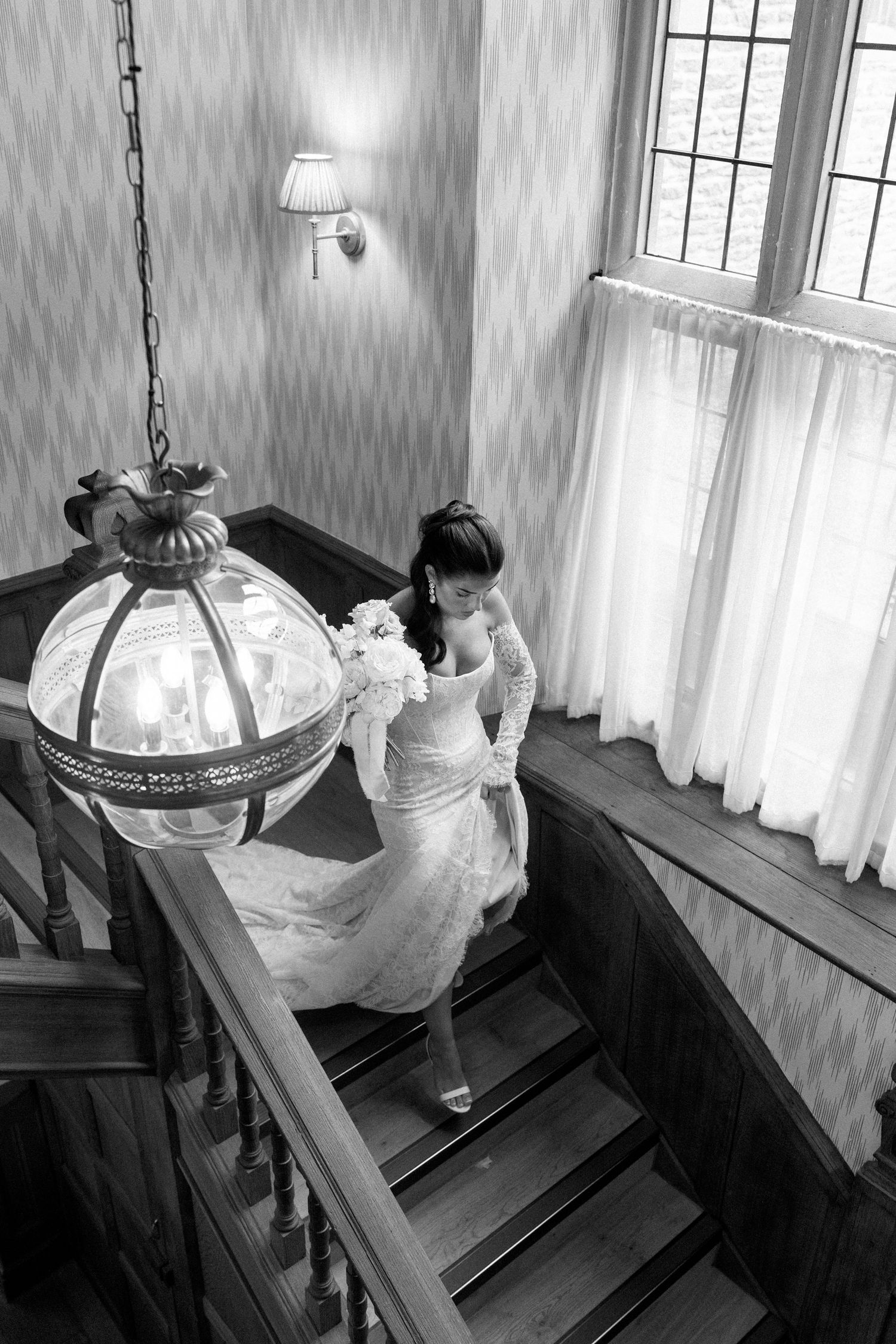 Black and white aerial view of bride in lace gown descending wooden staircase with bouquet.