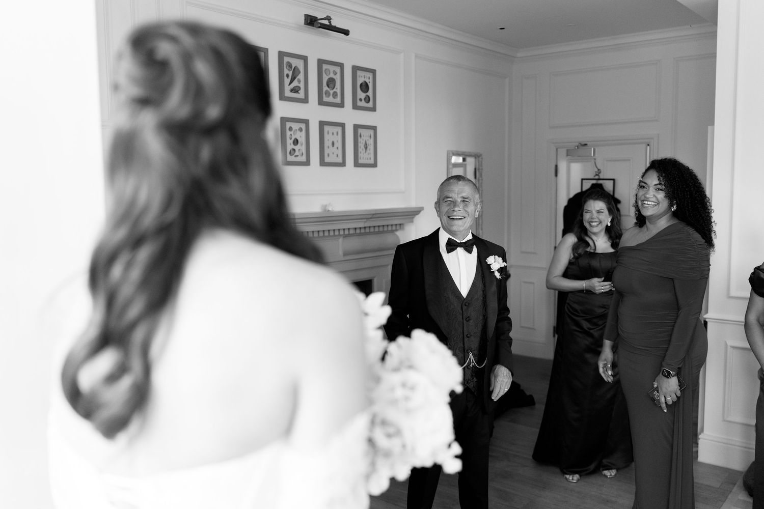 Bride in lace gown meets family members in elegant white room during wedding preparations.