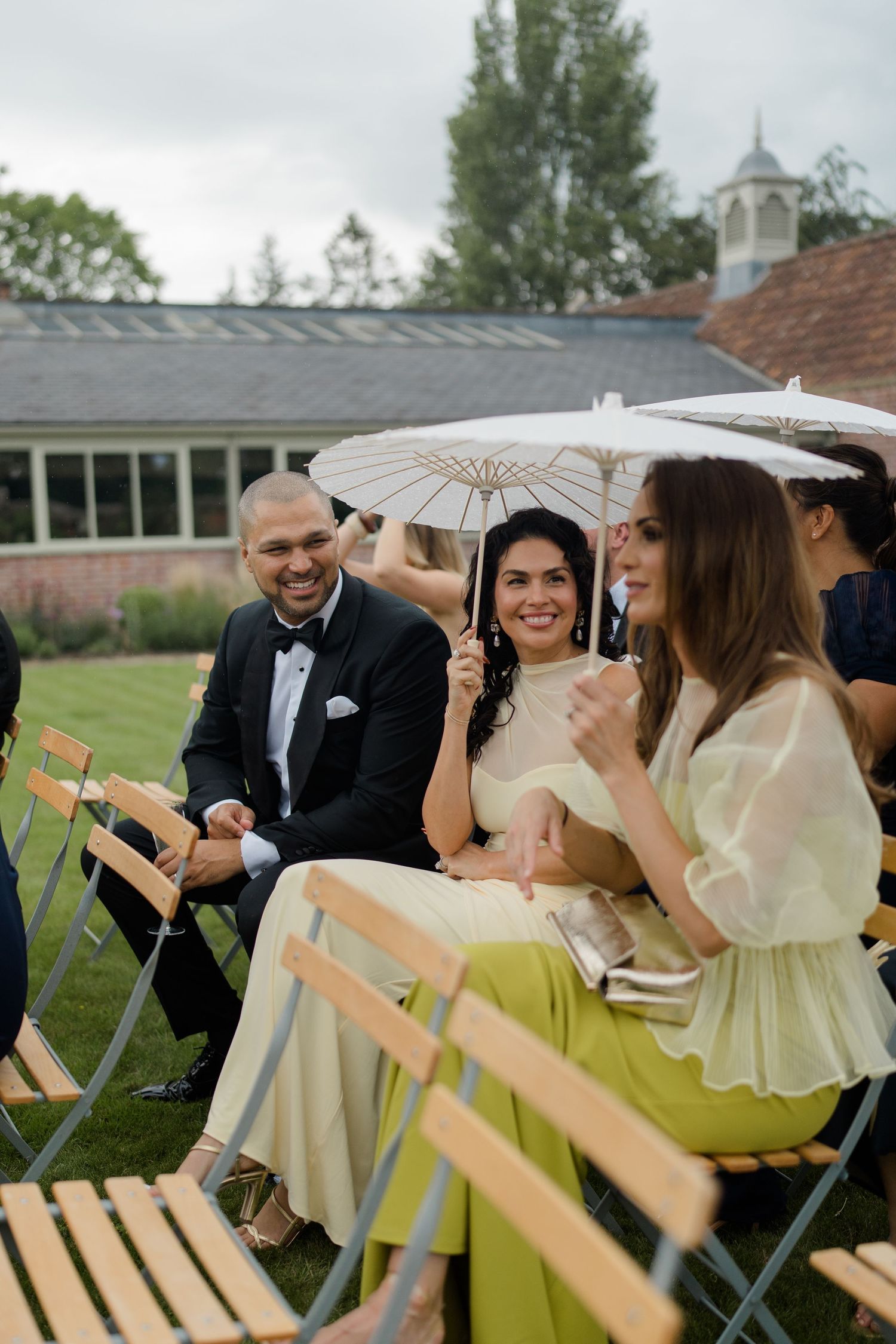 Wedding guests seated outdoors, smiling and holding parasols during a garden ceremony in formal wear.