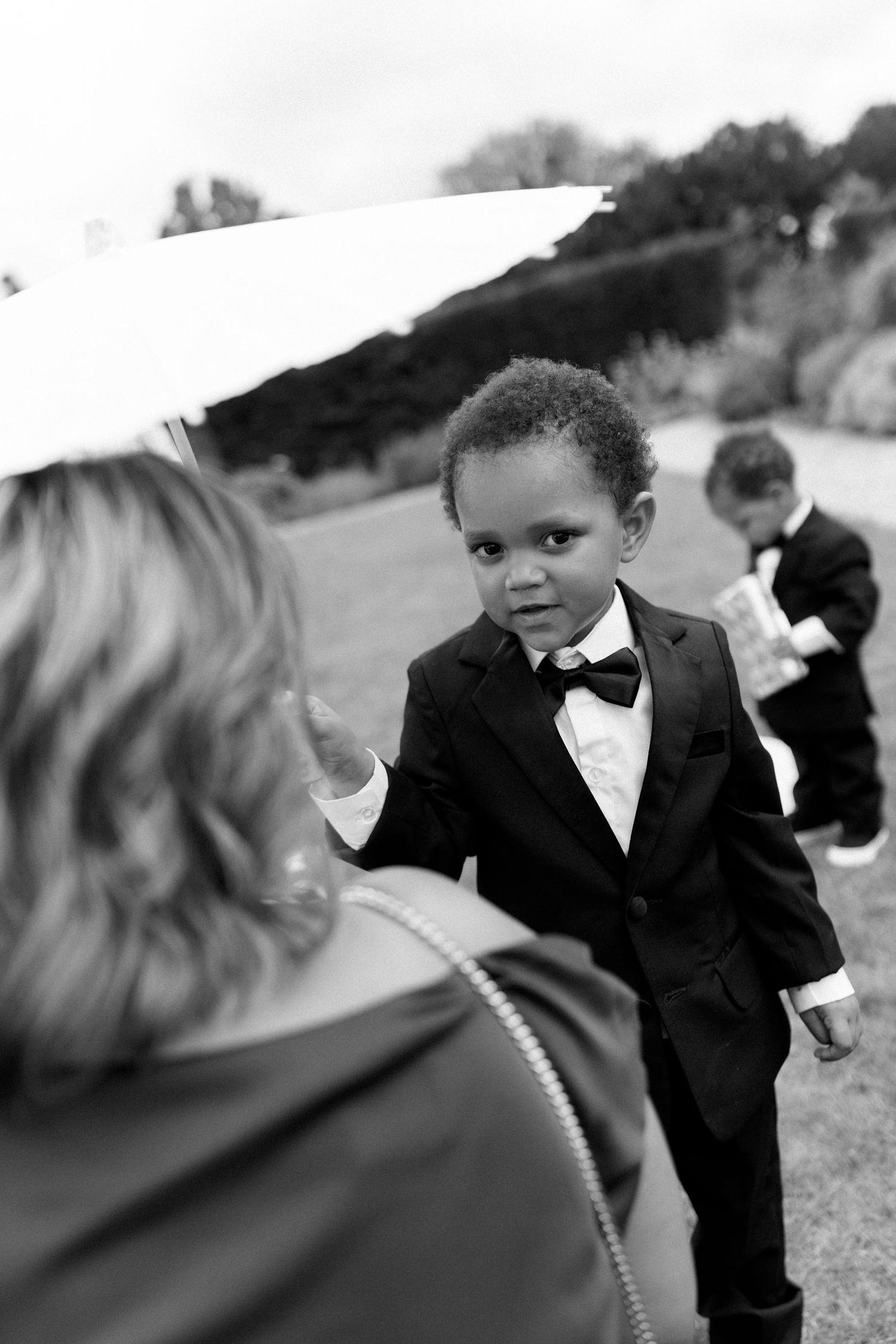 Young ring bearer in black tuxedo and bow tie reaches toward guest at outdoor wedding.
