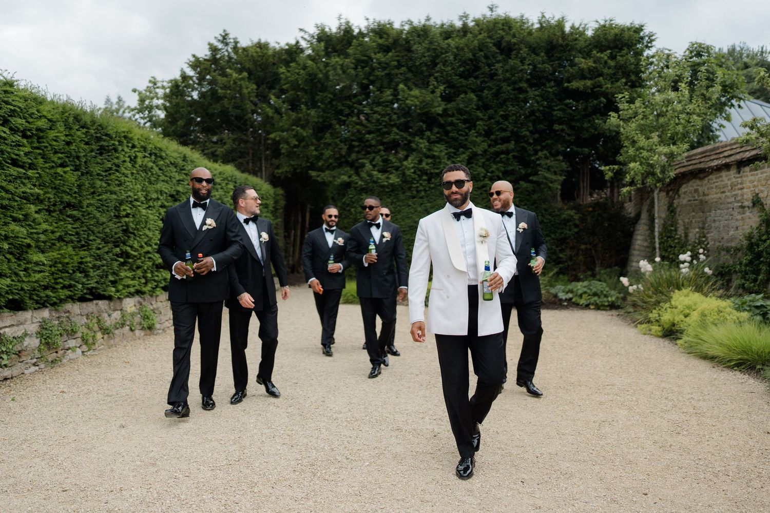 Stylish groom in white tuxedo walks confidently with groomsmen in black suits through garden.