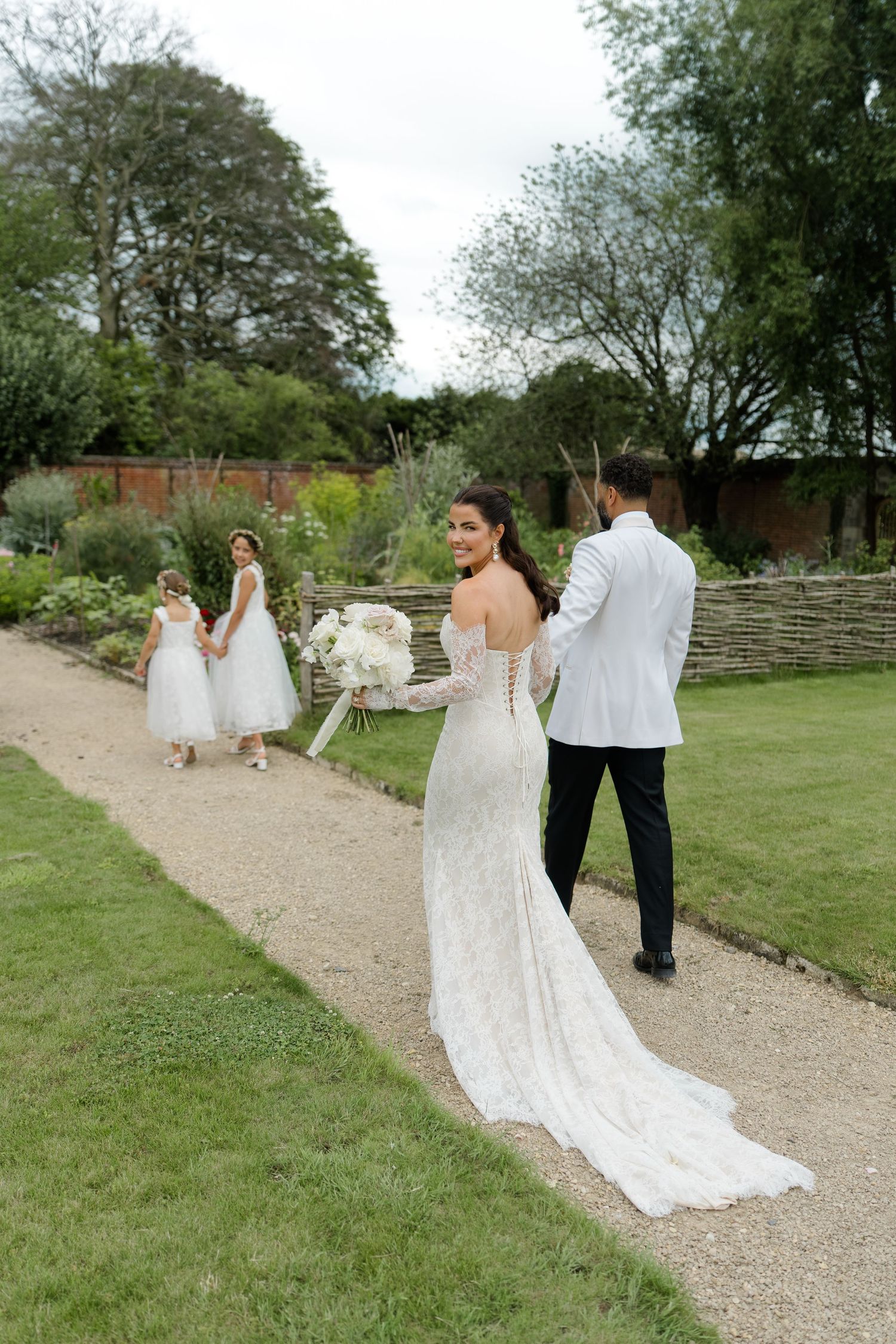A bride in a lace gown with train walks a garden path with her groom and flower girls.