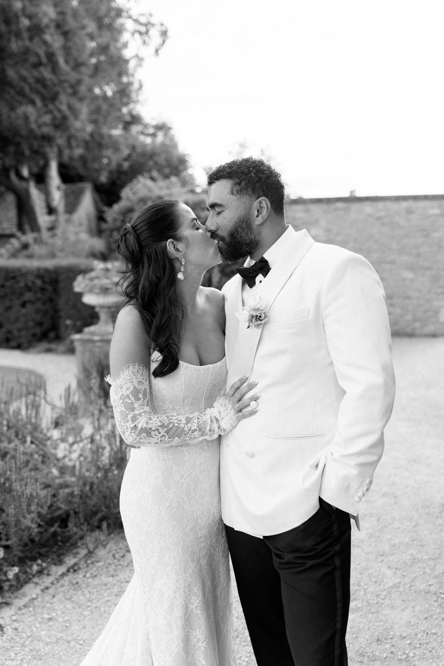 A black and white photo of a bride and groom sharing a kiss in a formal garden setting.