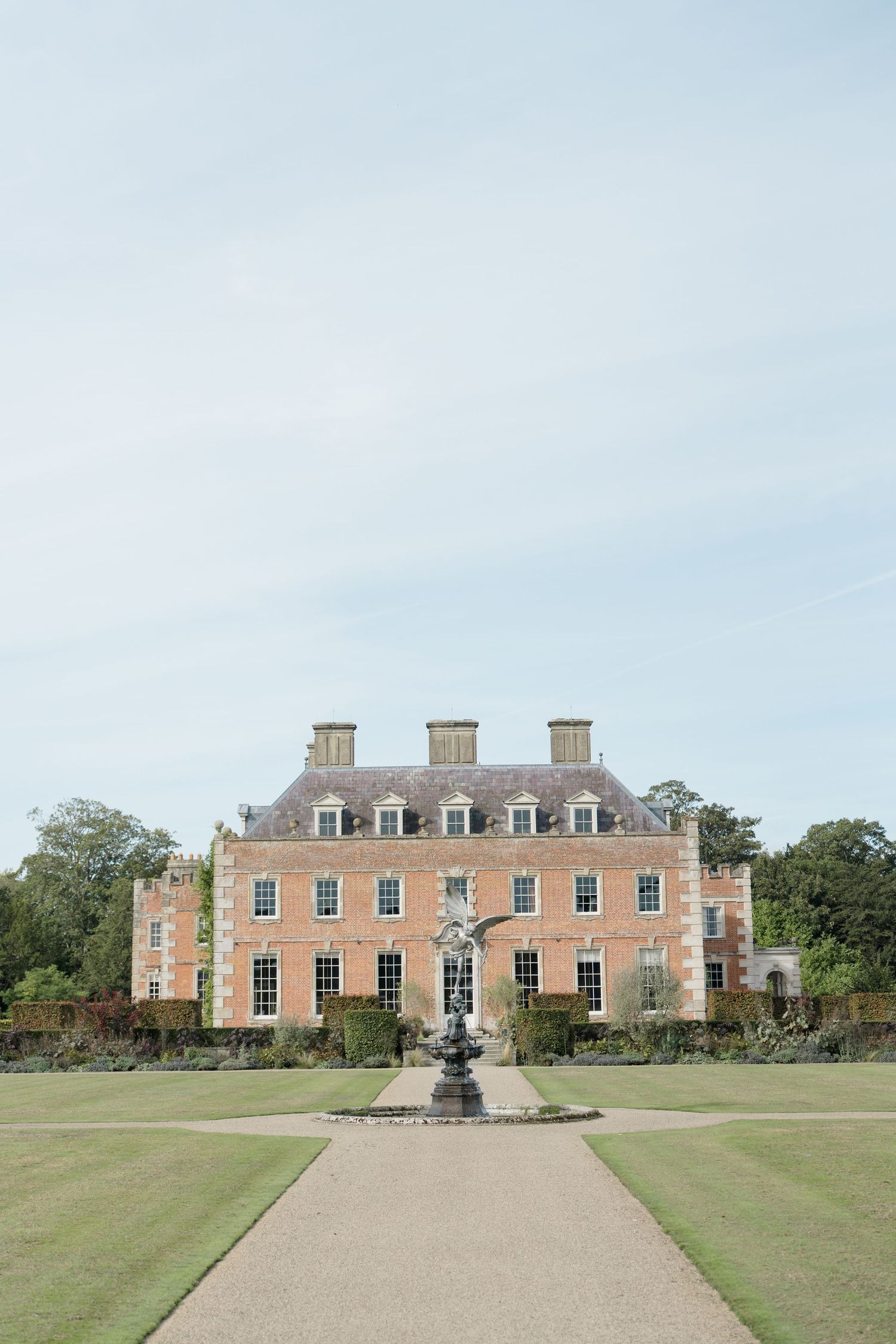 Grand Georgian red brick manor house with ornate fountain centerpiece, formal gardens, and gravel pathway under a pale blue sky.