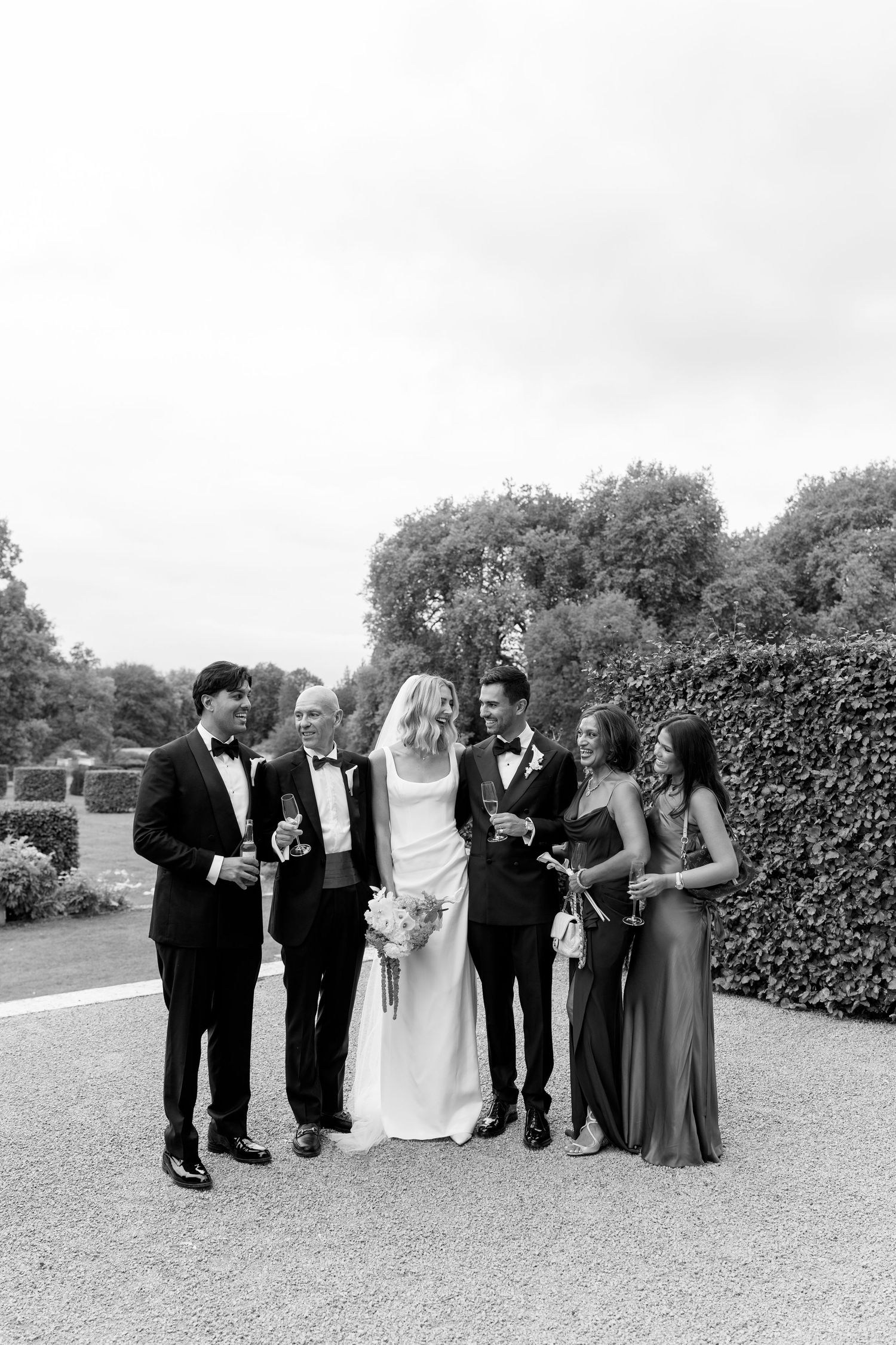 Bride and groom pose with wedding party in black and white photo outdoors among lush hedges.