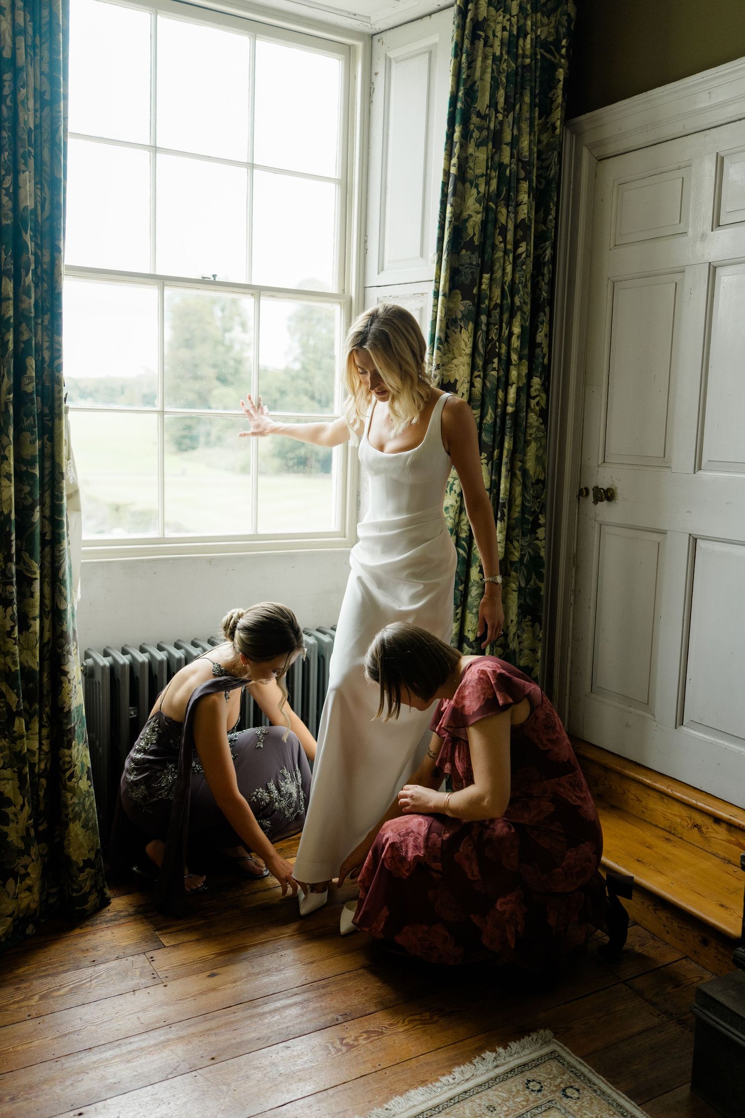 Two bridesmaids help a bride into her white wedding shoes by a large window with garden views.