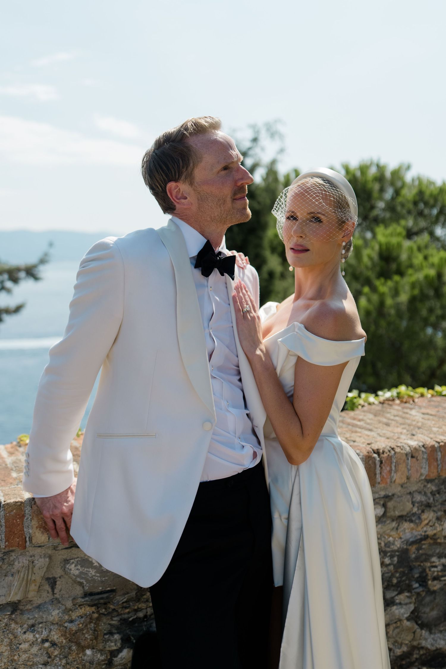 Stylish bride in birdcage veil embracing groom in white tuxedo at scenic outdoor venue.