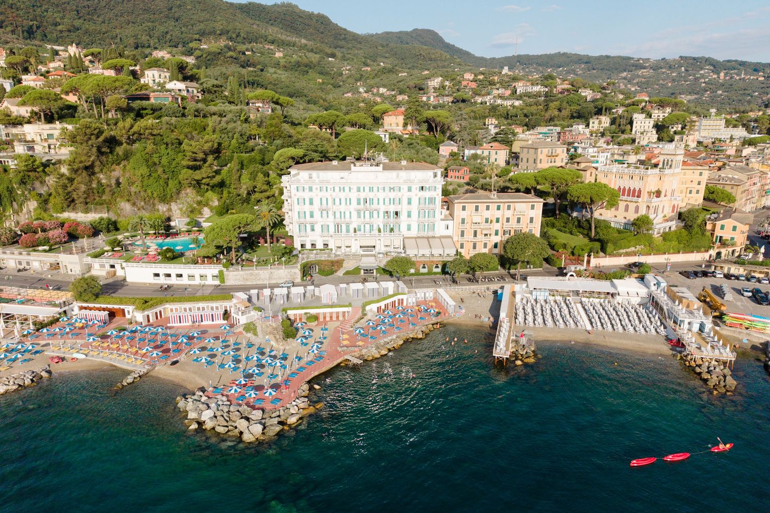 Aerial view of a luxurious beachfront hotel in Italy with colorful beach umbrellas, rocky coastline, and lush green hills.