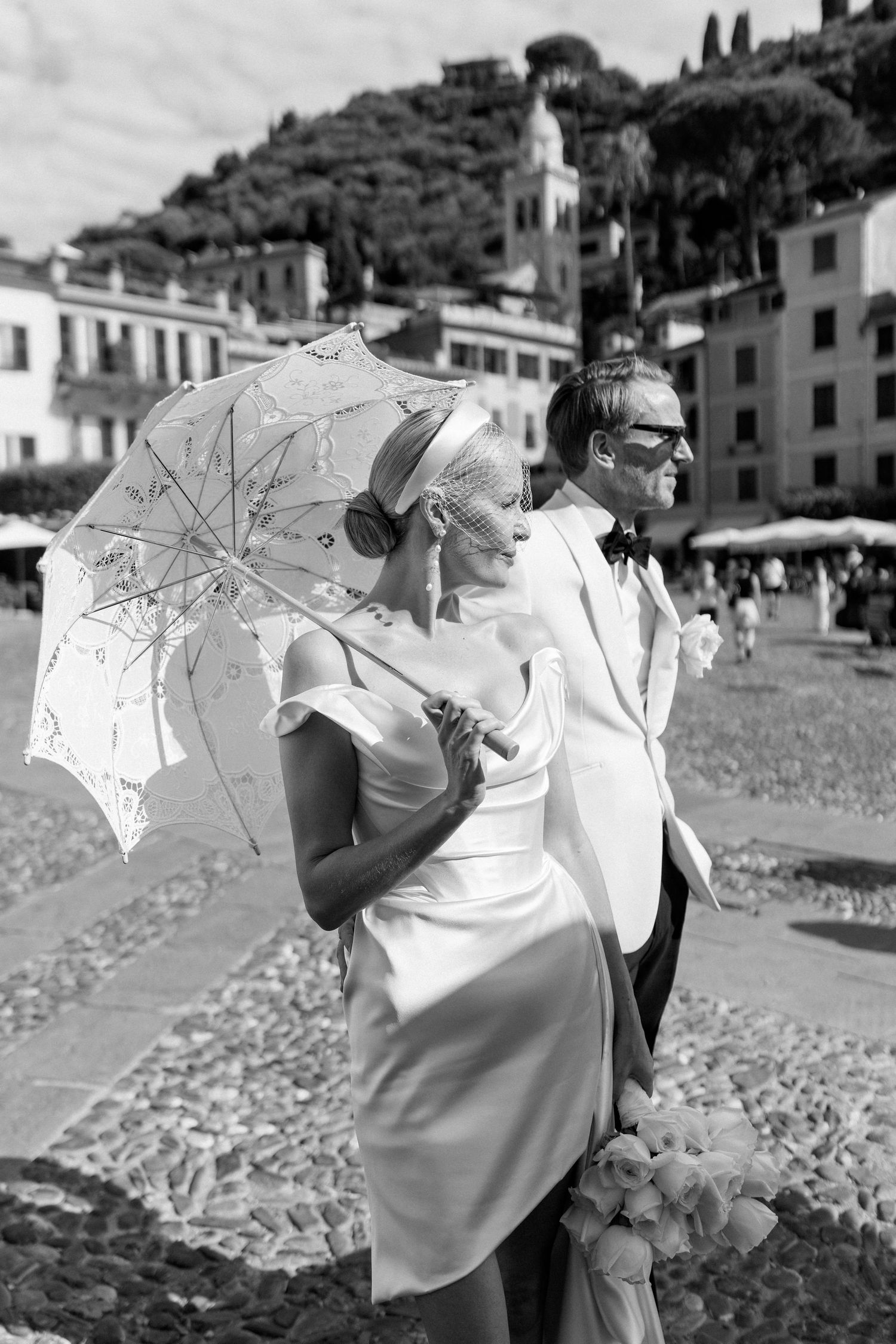 Black and white photo of a bride and groom with a lace parasol in a charming Italian coastal piazza.