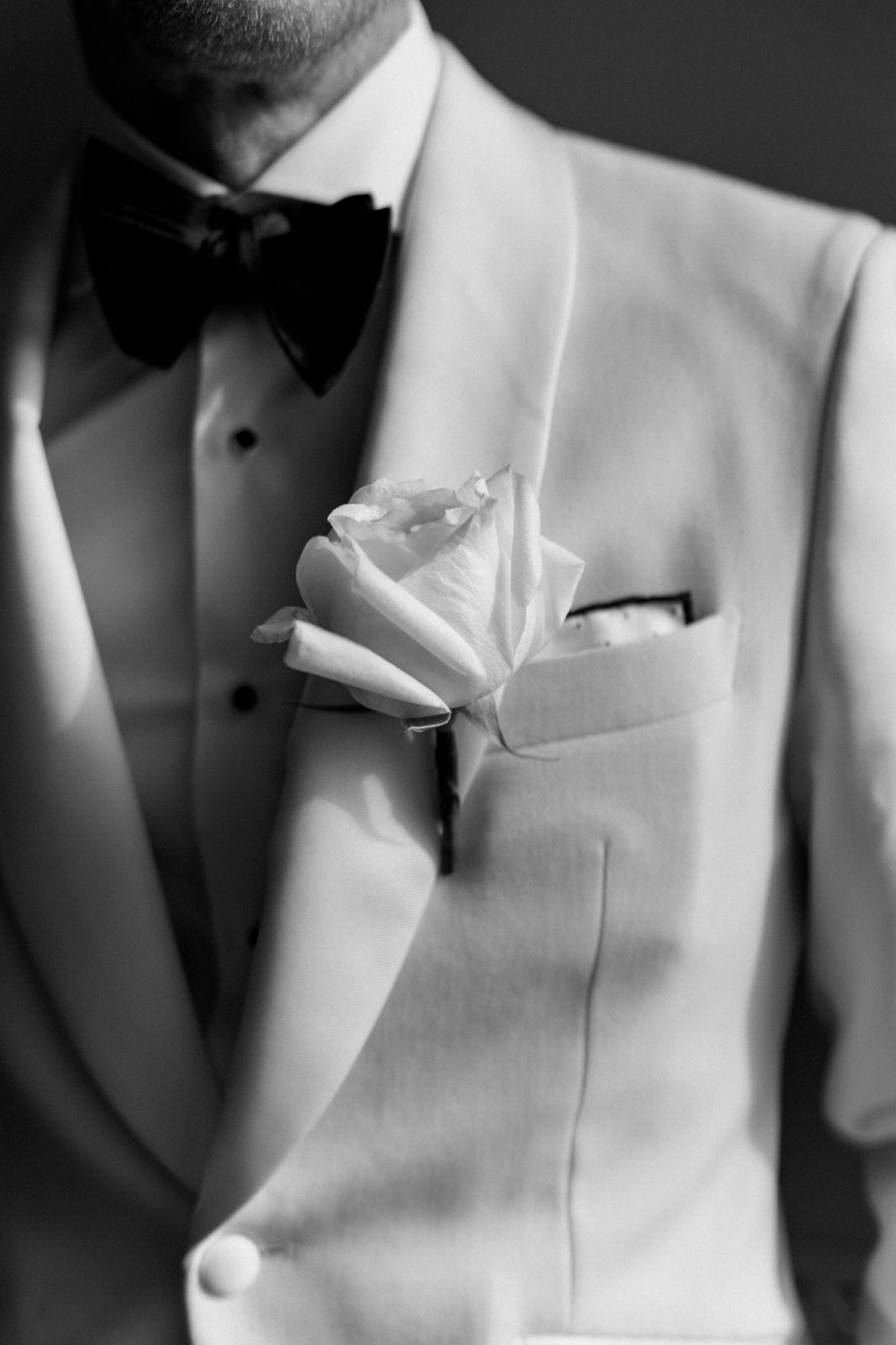 Black and white close-up of groom in white tuxedo with black bow tie and white rose boutonniere.