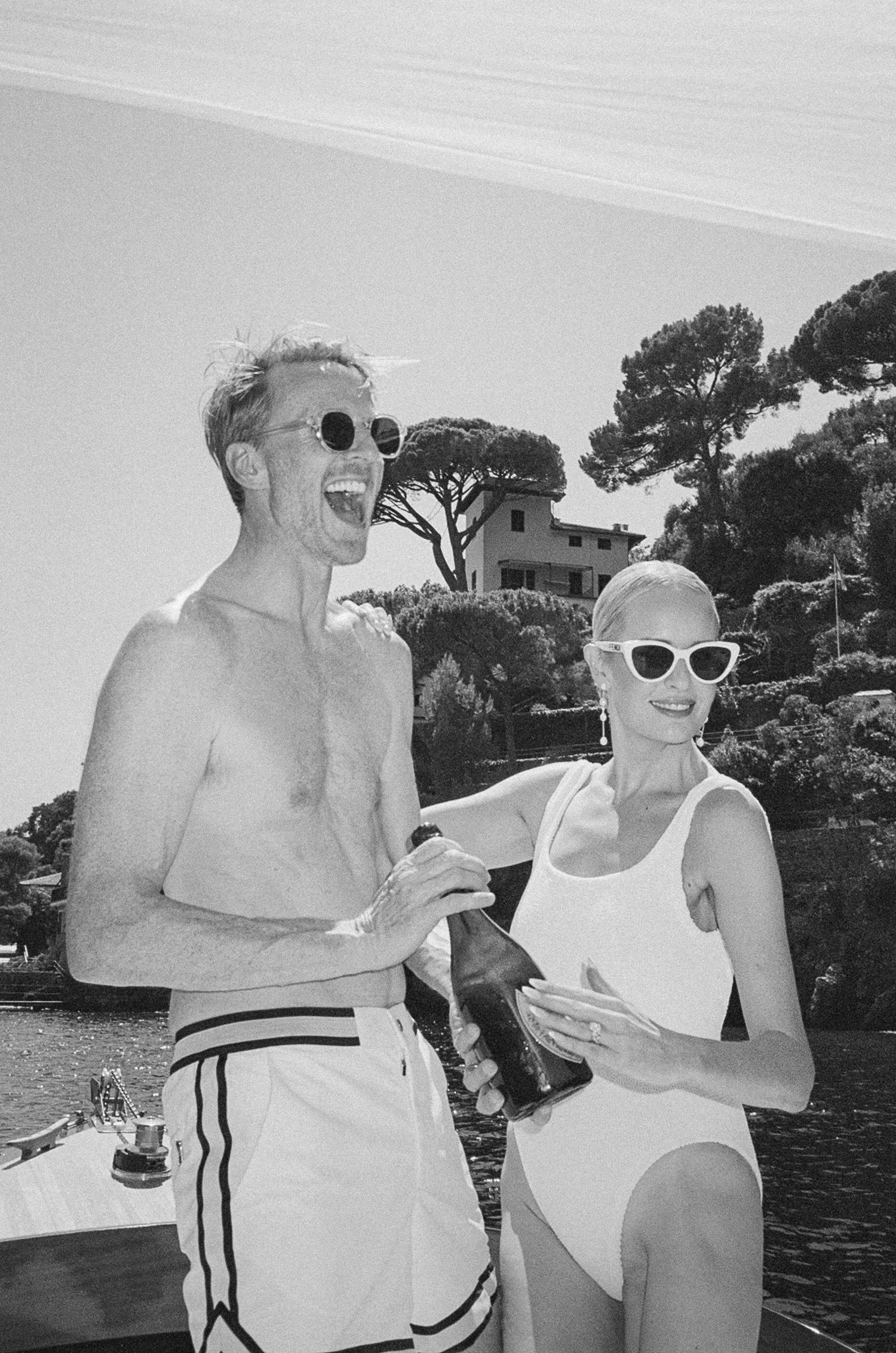 Couple in swimwear laughing with champagne bottle on boat in black and white photo.