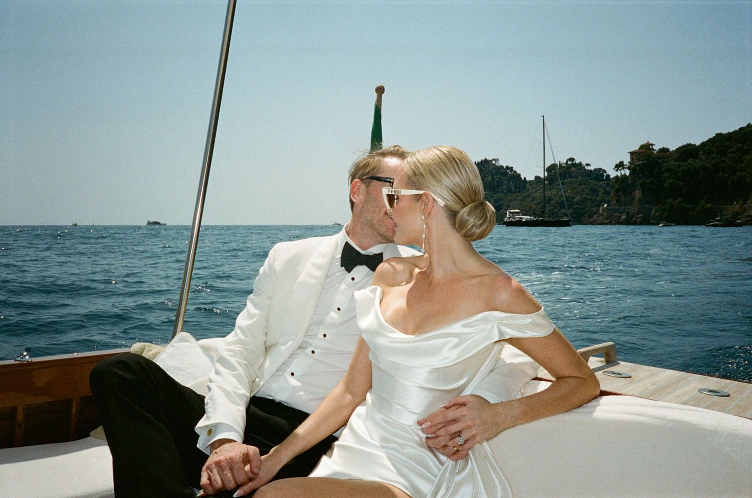 A newlywed couple in wedding attire shares a romantic kiss on a sailboat on a sunny day.