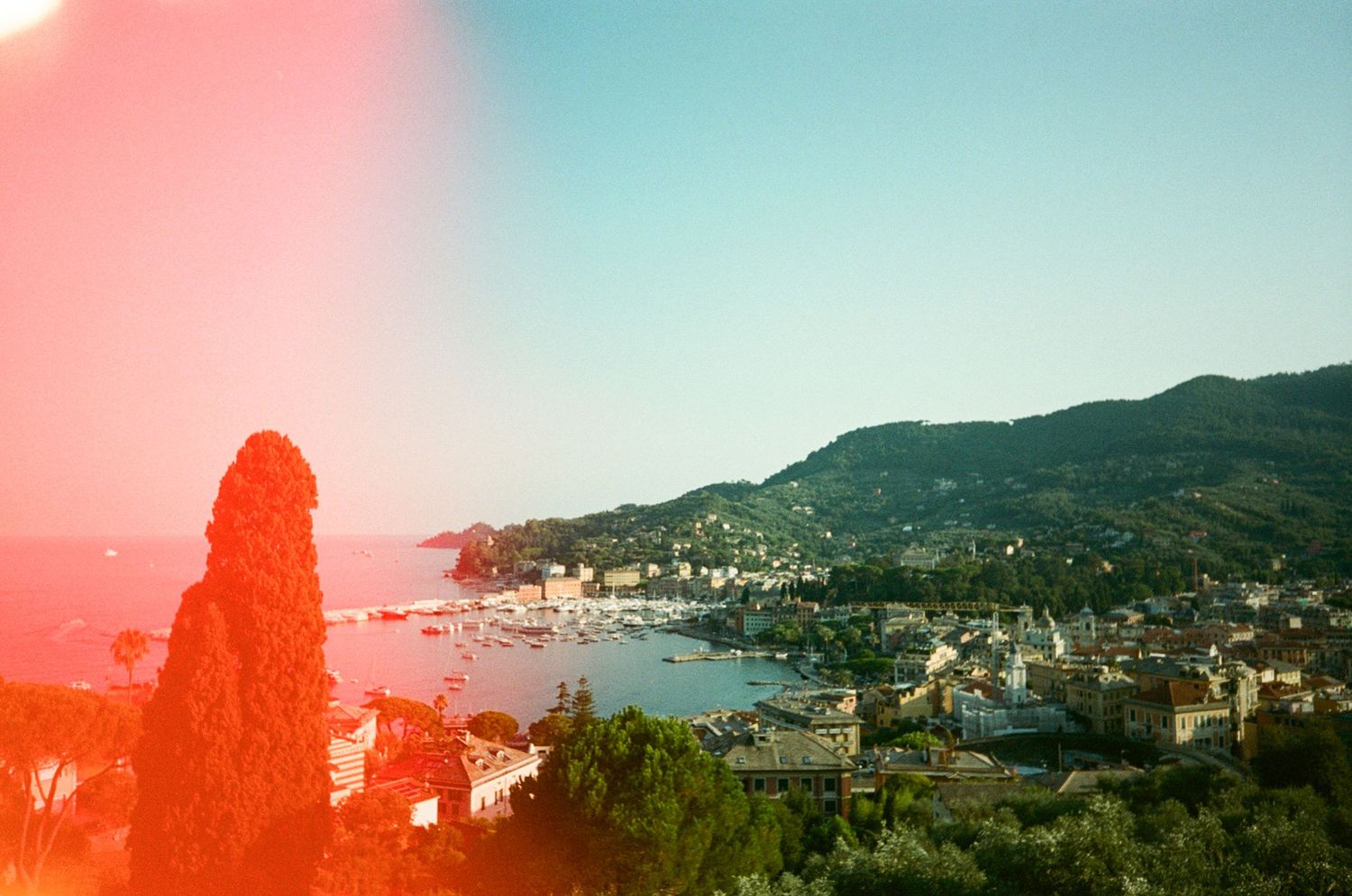 Aerial film photo of Italian coastal town with harbor, lush hills, and colorful light leak overlay.