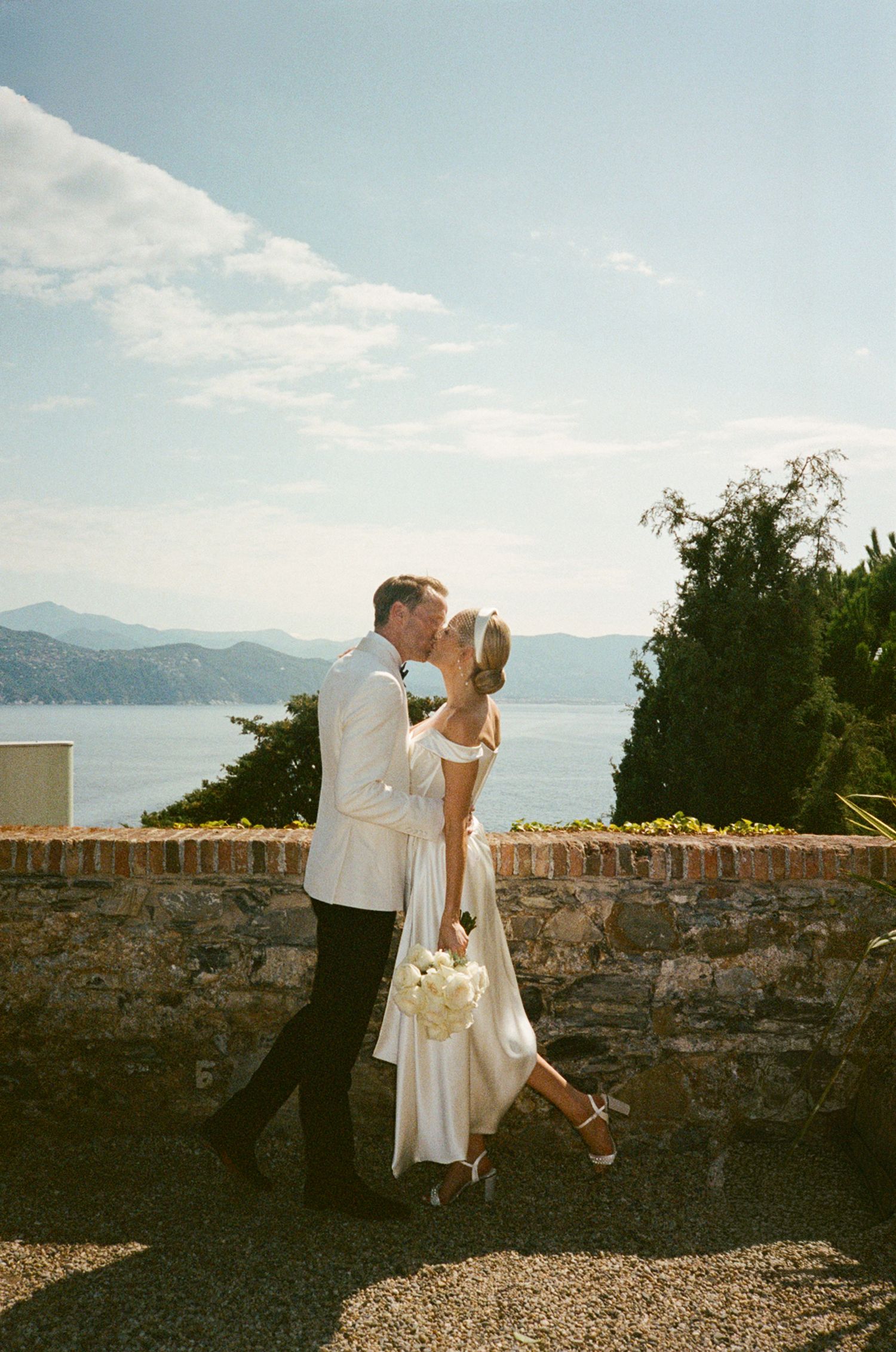 Bride and groom share a romantic kiss by a stone wall with a stunning lake and mountain backdrop on their wedding day.