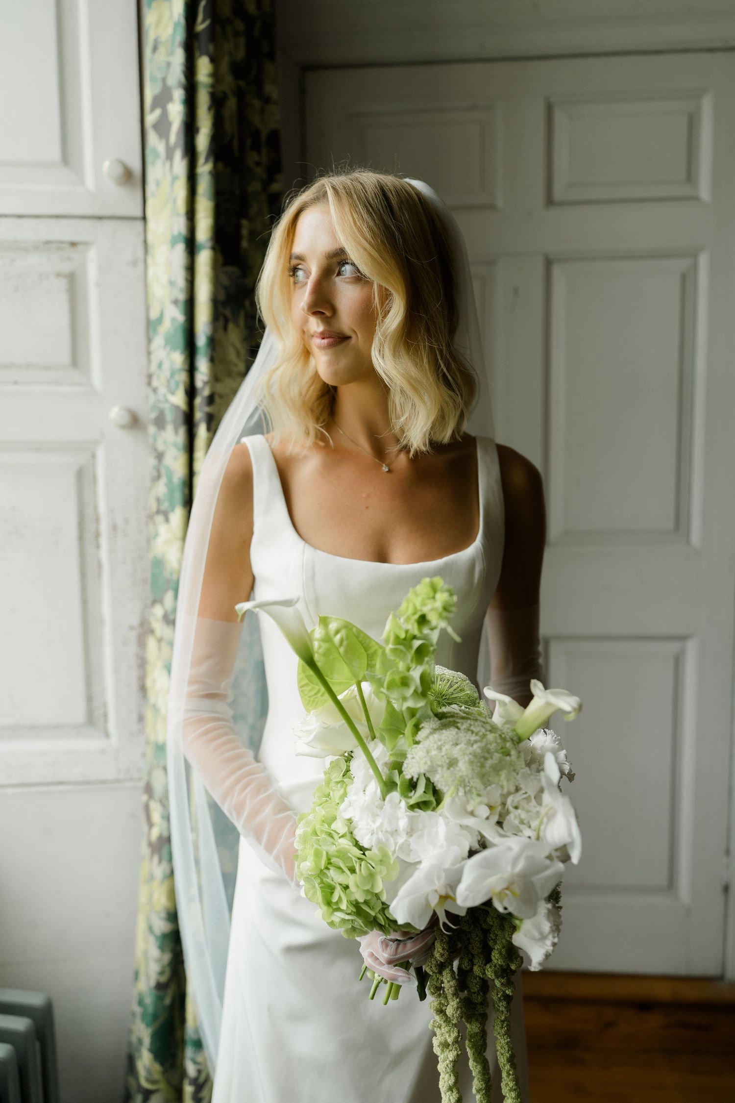 Bride in white gown and veil holds green and white floral bouquet, gazing toward window in elegant room.