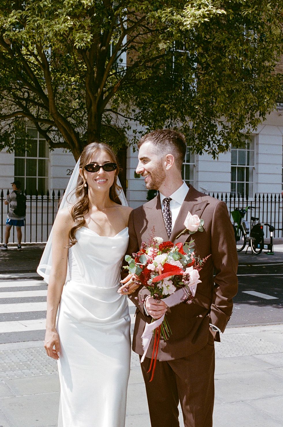 Smiling newlyweds in white gown and brown suit pose outdoors holding red wedding bouquet.
