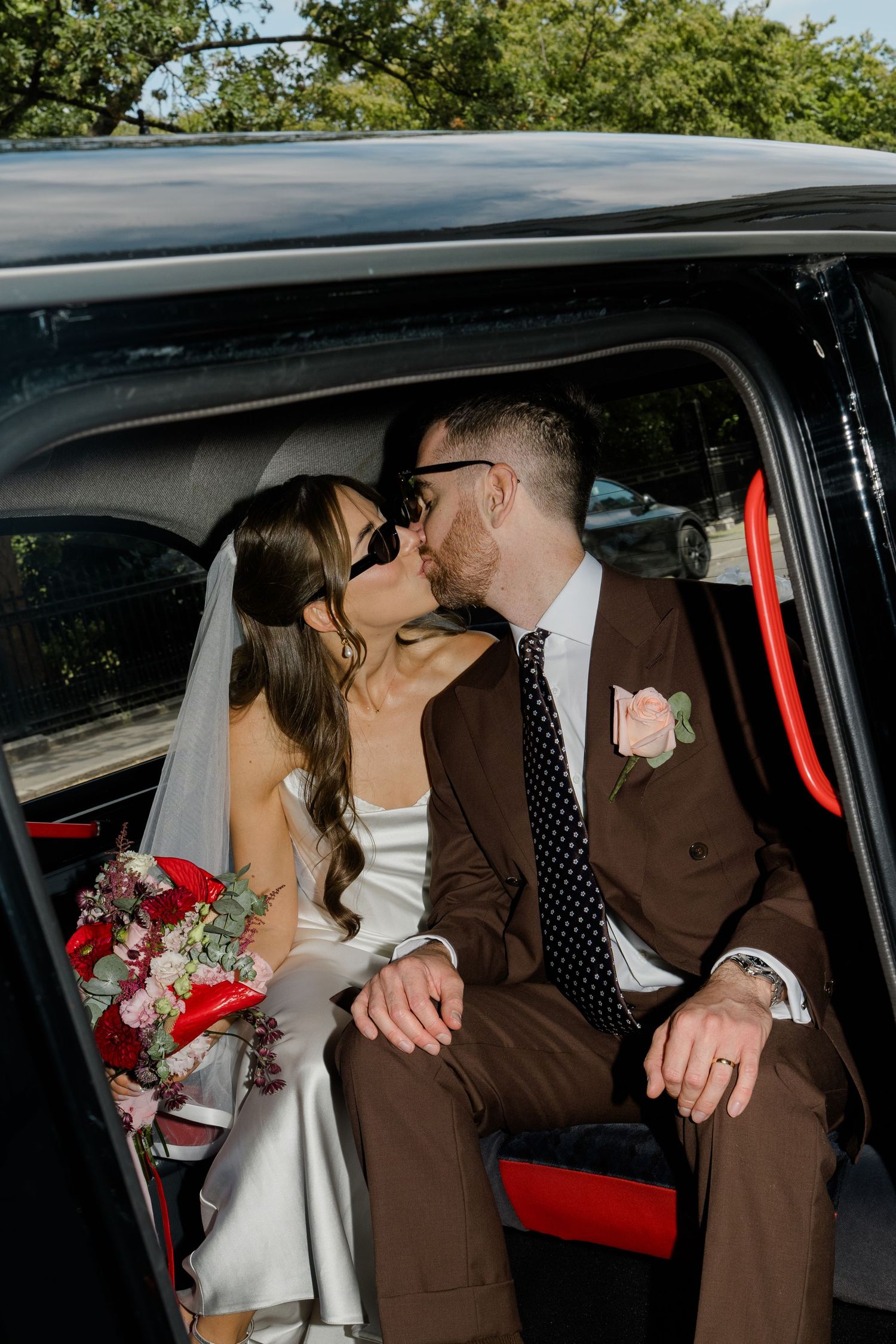 Bride and groom share a kiss in a black London taxi cab, bride holding red and pink bouquet.