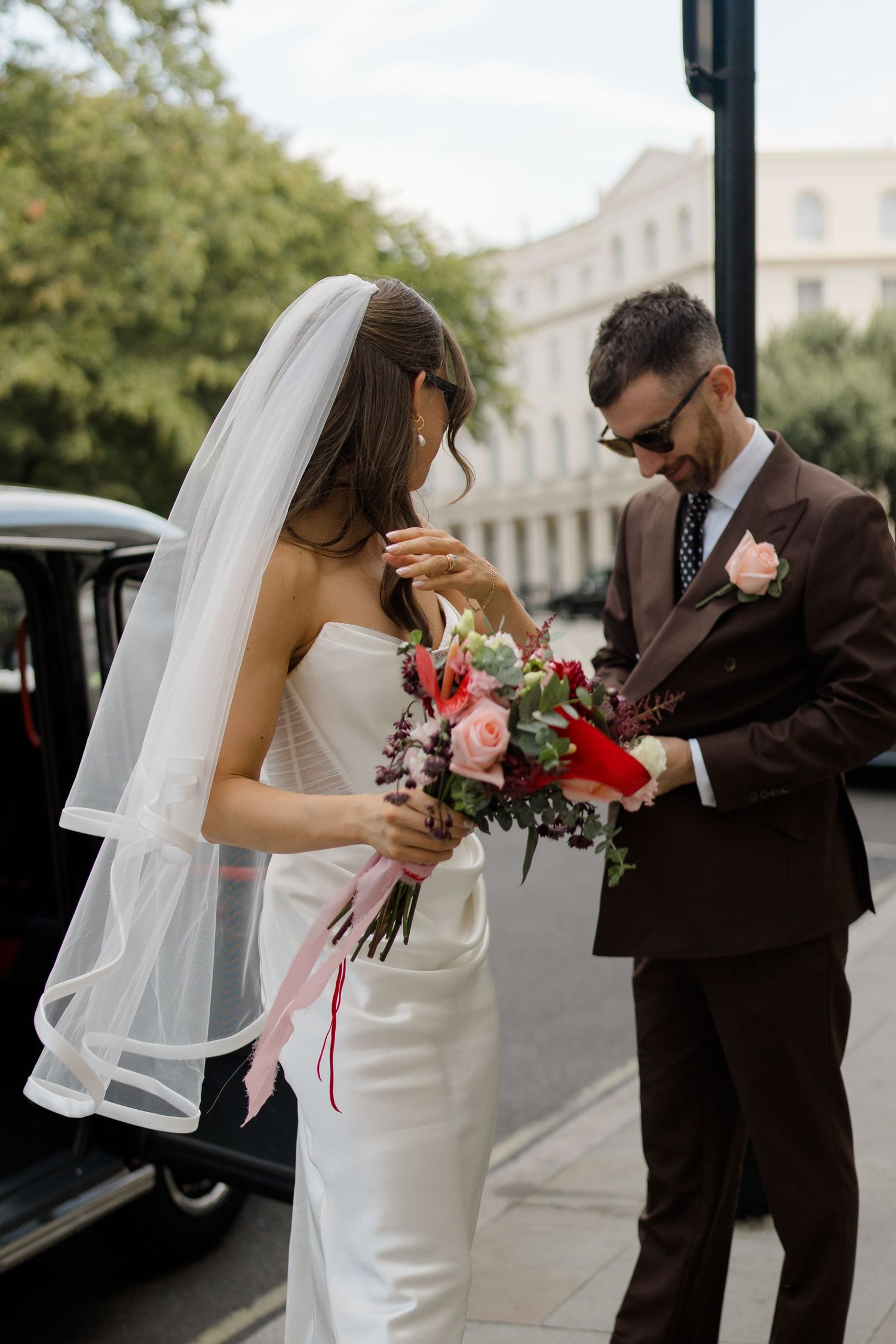 Bride and groom in sunglasses admire wedding bouquet of red and pink flowers beside black car.