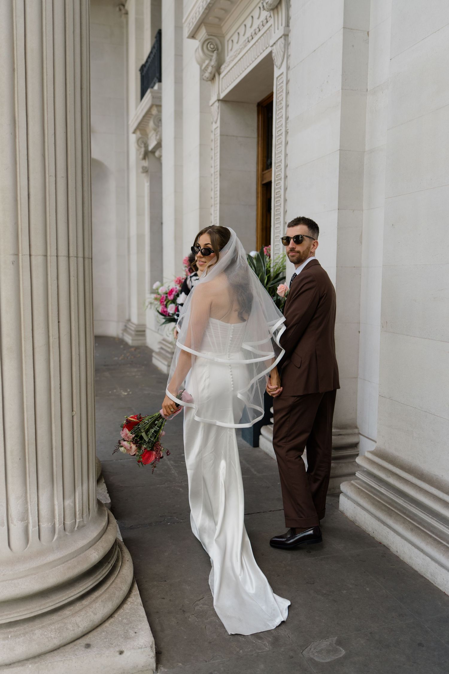 Bride in white gown with veil and groom in brown suit posing between grand columns with bouquet.