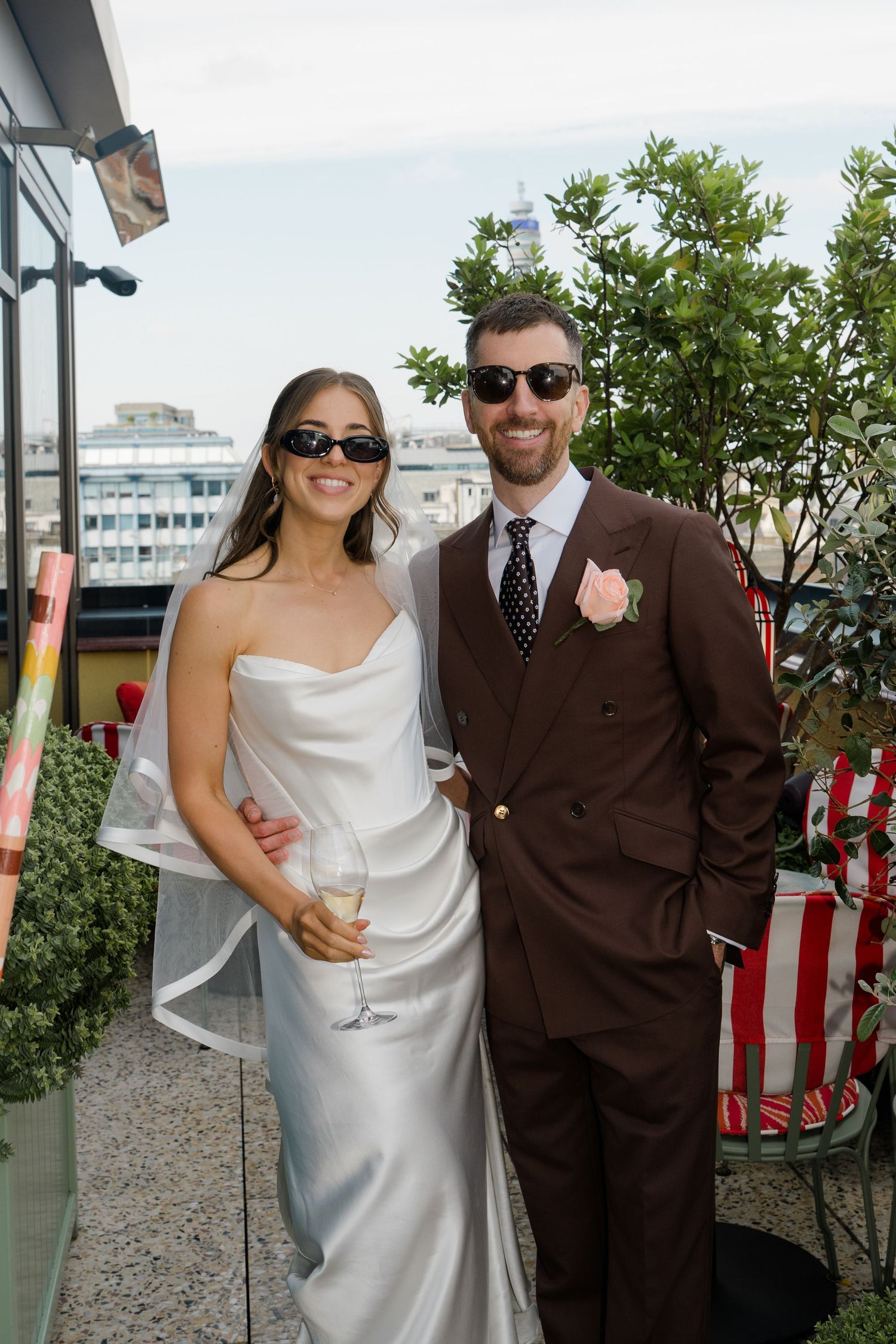 Bride in silver gown and groom in brown suit wearing sunglasses on a rooftop wedding celebration.